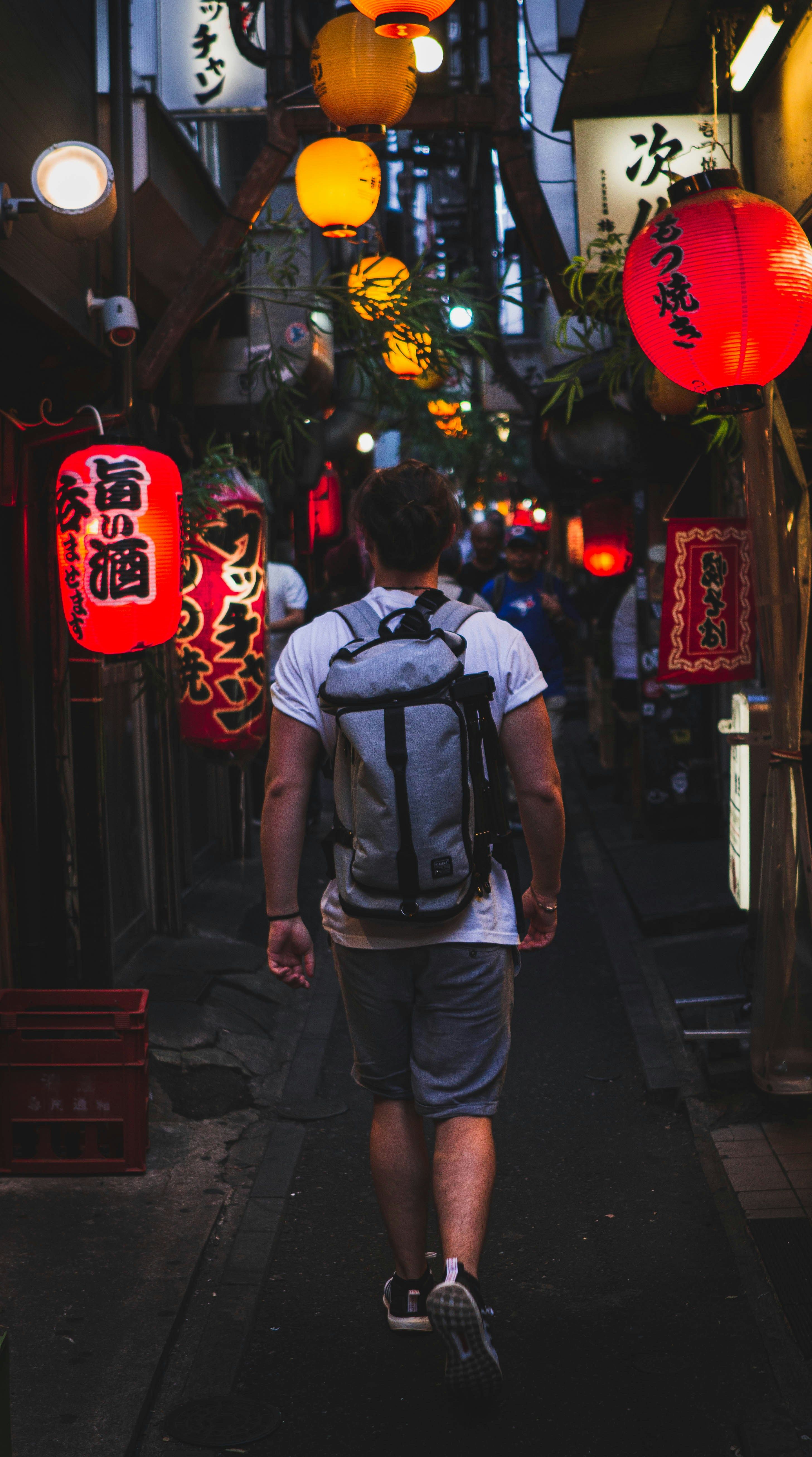 Rear view of a person with a backpack walking through a narrow Japanese alley lit by red and yellow lanterns.
