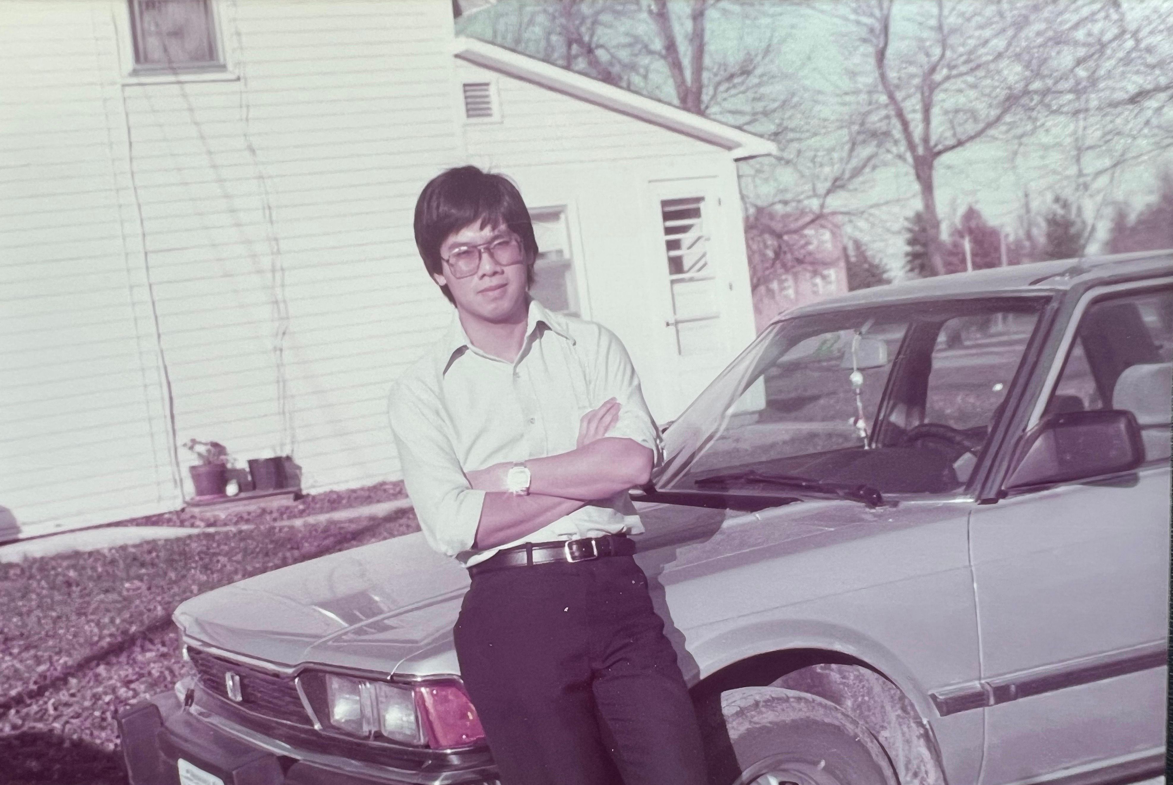 A portrait of a man standing in front of a silver car with a white house in the background