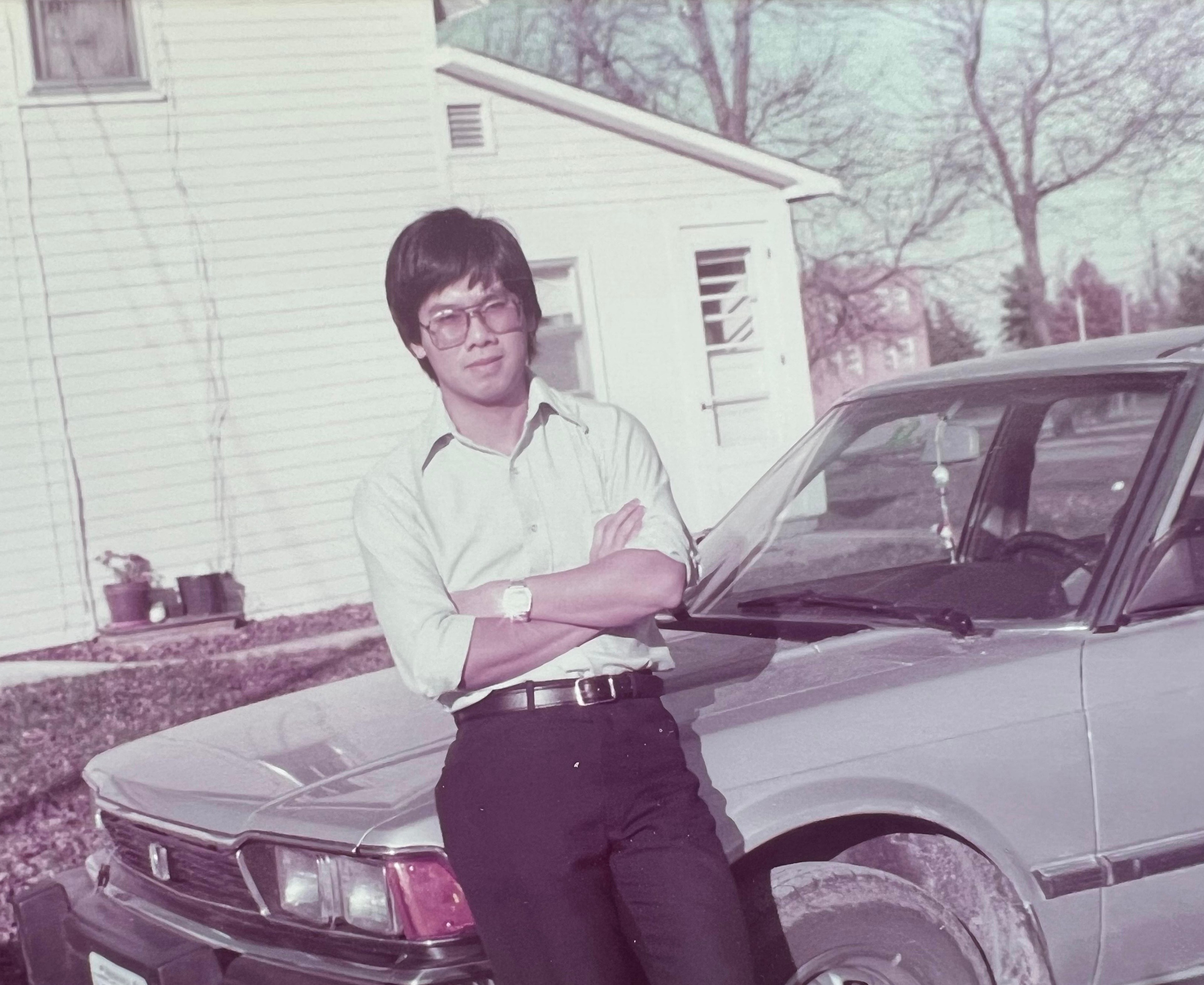 A portrait of a man standing in front of a silver car with a white house in the background