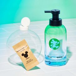 Disney & Blueland bottle with Mickey Mouse logo on white tile next to table in compostable wrapper and 2 naked tablets on bubble with green background
