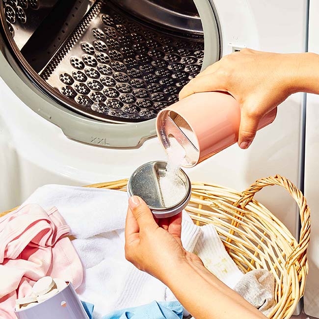 Forever tin pouring powder into lid next to washing machine over basket of laundry