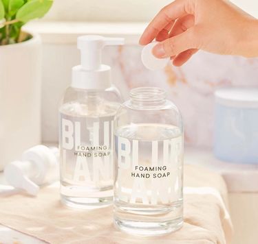 Hand Soap Duo: 2 glass bottles on kitchen counter with hand dropping tablet into open soap bottle of water