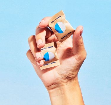 Hand holding two Blueland Glass + Mirror refill tablets in compostable wrappers against blue background