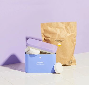 Toilet bowl cleaner starter set: refillable tablet tin with tablets sticking out on white tile and purple background and compostable refill pouch
