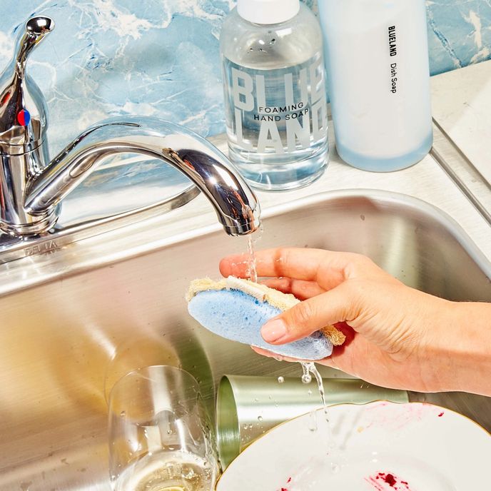 Hand holds scrub sponge under water running in sink of dirty dishes