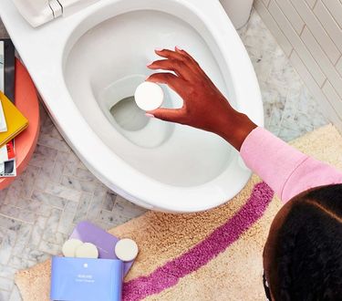 Hand drops toilet bowl cleaner tablet into toilet. Blueland toilet bowl cleaning tablets in purple tin next to toilet.