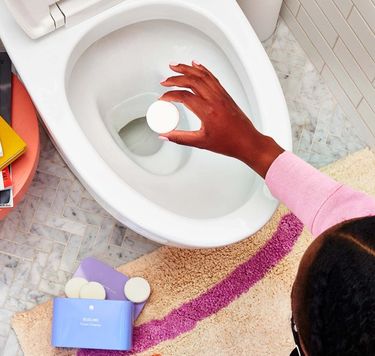 Hand drops toilet bowl cleaner tablet into toilet. Blueland toilet bowl cleaning tablets in purple tin next to toilet.