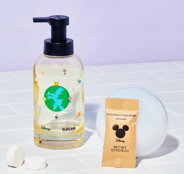 Disney & Blueland bottle with Daisy Duck logo on white tile next to table in compostable wrapper and 2 naked tablets on bubble with purple background