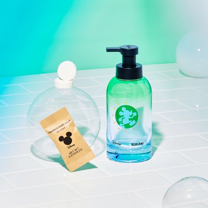 Disney & Blueland bottle with Mickey Mouse logo on white tile next to table in compostable wrapper and 2 naked tablets on bubble with green background