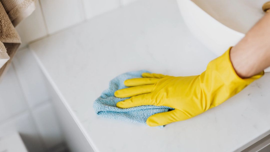 yellow rubber gloved hand cleaning a white counter with blue cleaning cloth