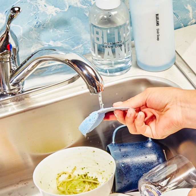 Hand holds pop-up sponge under water running in sink of dirty dishes