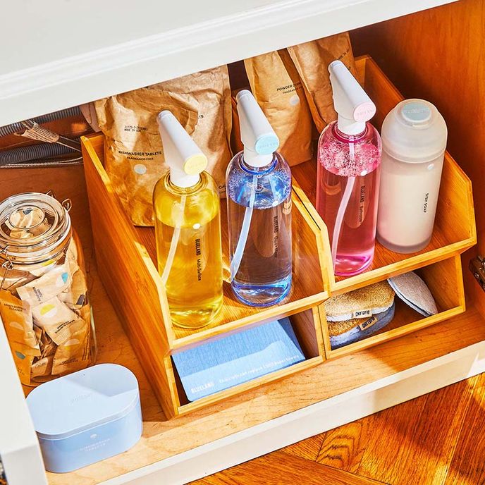 Assortment of Blueland cleaners under sink on organized wooden shelf holders. Combination of spray bottles, sponges, dish powder, and refills.