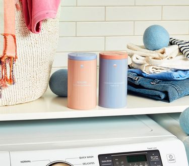 Laundry Essentials Kit: 2 refillable Forever tins on laundry shelf above laundry machine with folded clothes and Laundry Balls behind