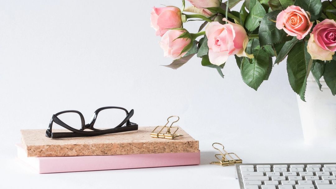 pink roses in a vase, keyboard in foreground and notebooks and glasses stacked on top of eachother