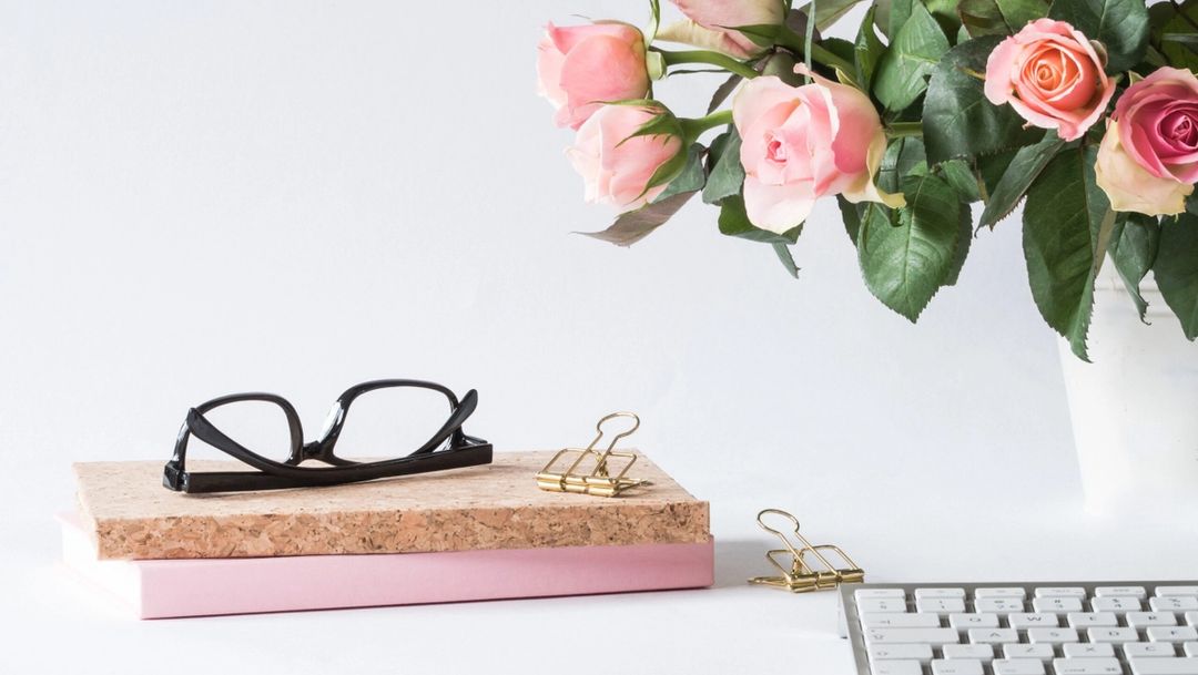 pink roses in a vase, keyboard in foreground and notebooks and glasses stacked on top of eachother
