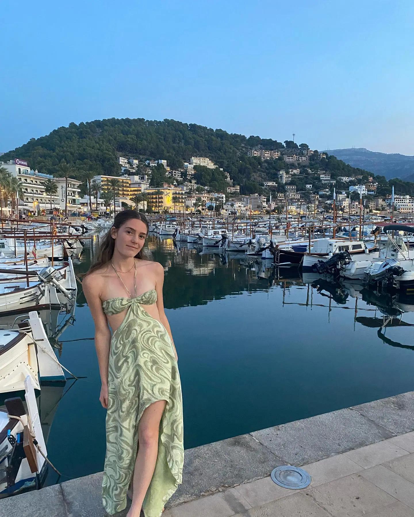 Person seated by a marina, with boats docked in the water behind them, wearing a light green dress.