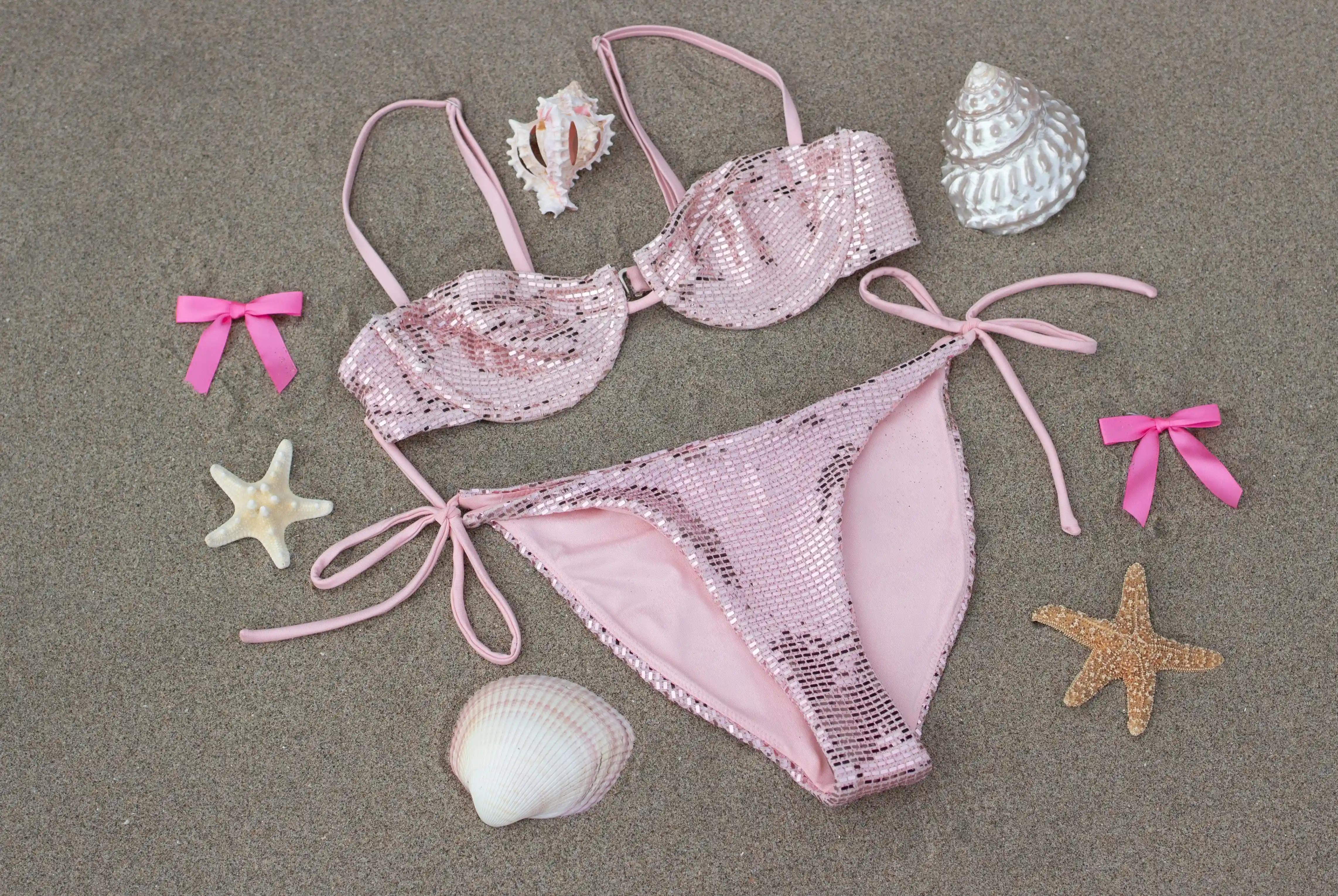 Pink sequin bikini set laid out on sand, surrounded by decorative seashells, starfish, and pink ribbon bows.