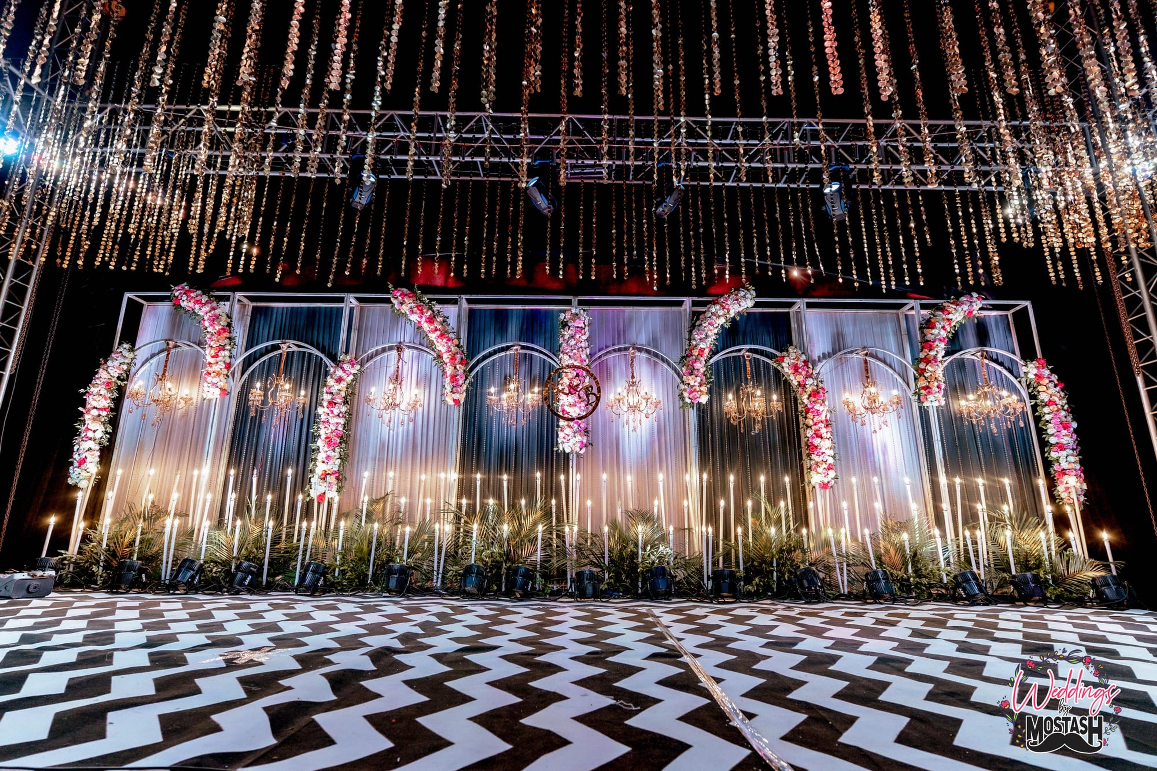 A glittering reception stage with a cascading fairy-light curtain, floral arches set with crystal chandeliers and a chevron floor