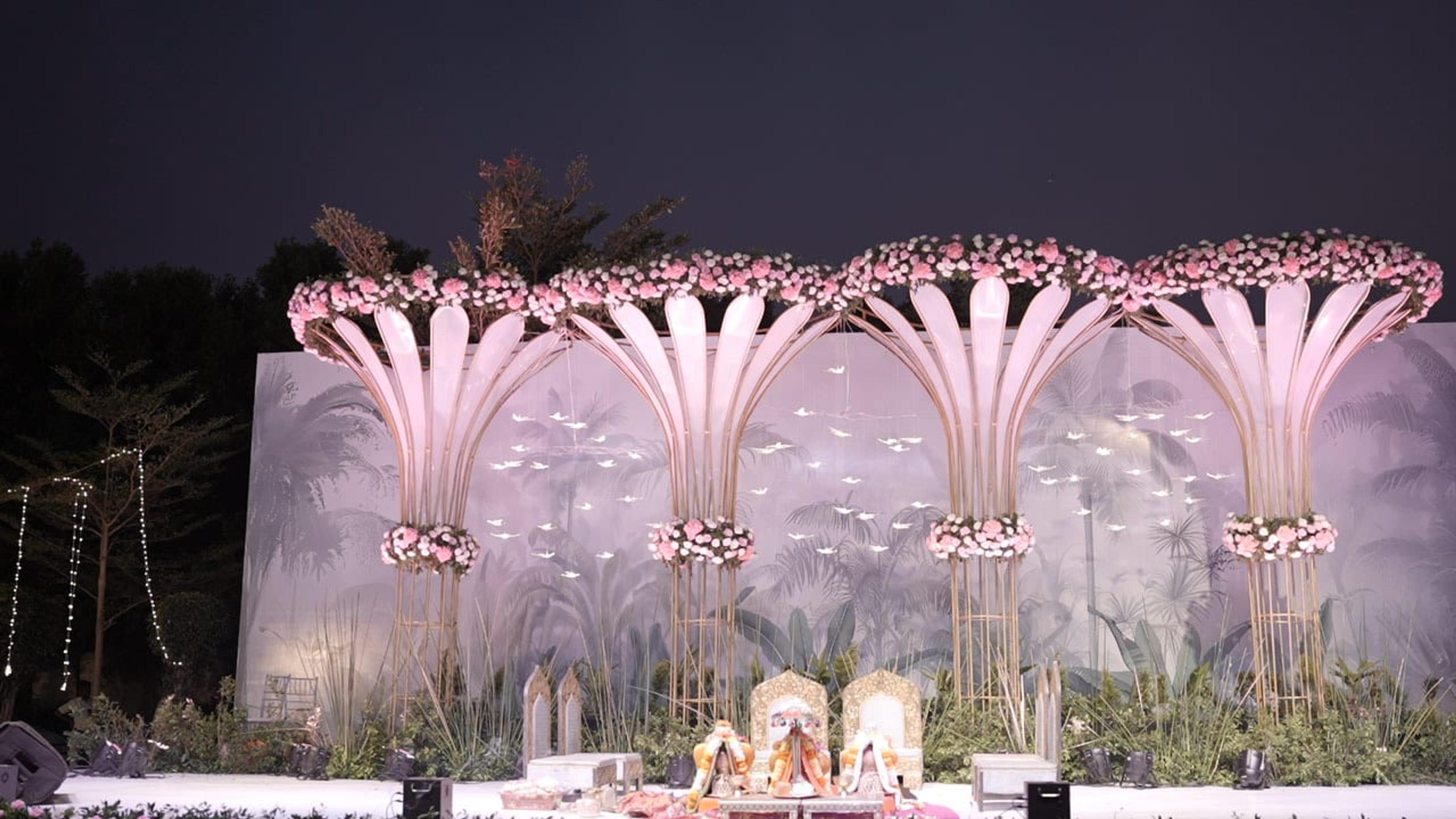 A grand outdoor stage backed by towering illuminated blush-pink palm-fan installations rising into the night sky