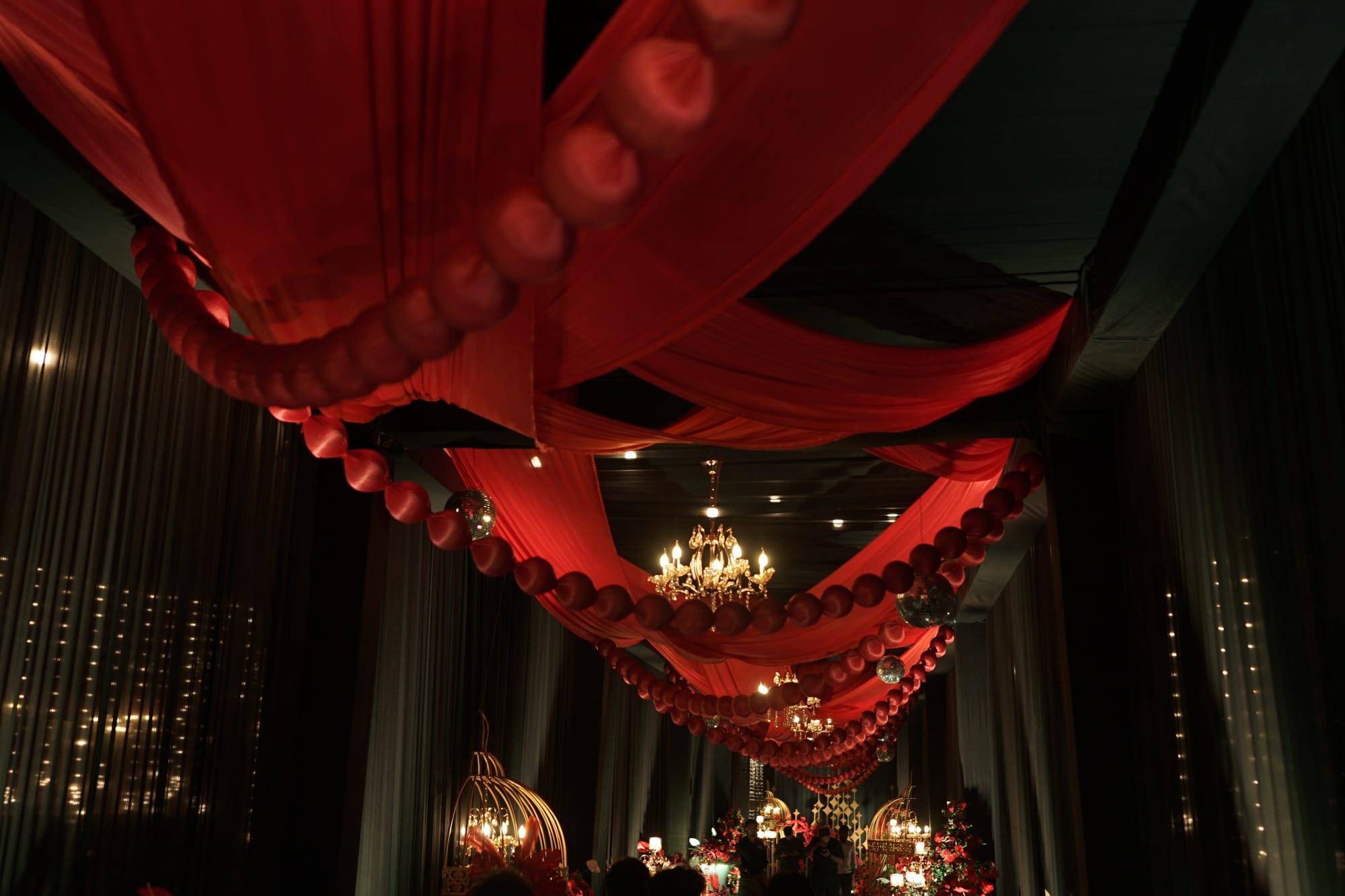 A dramatic red-draped ballroom ceiling canopy hung with crimson lanterns and warm crystal chandeliers