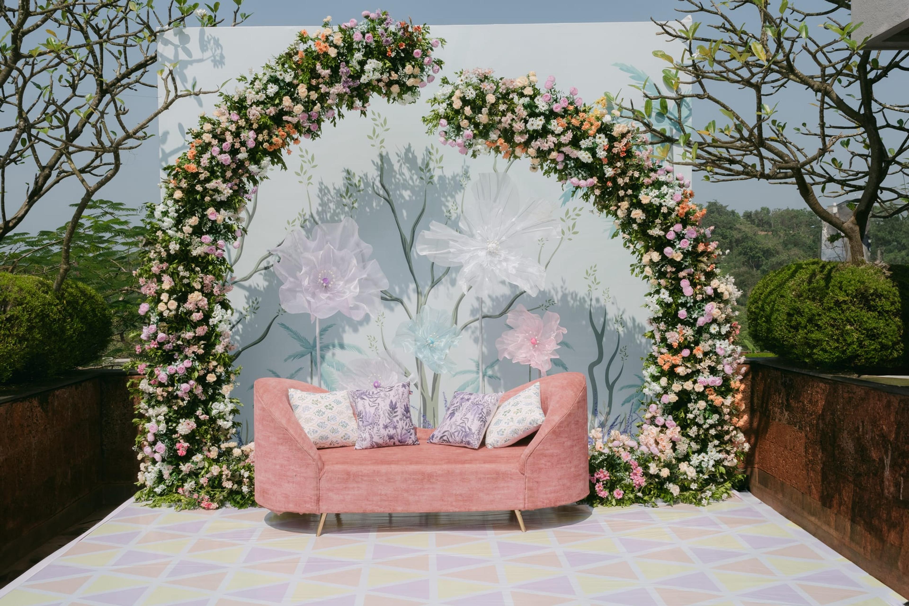 A blush velvet sofa on a hilltop mehndi stage framed by a pastel painted floral arch and flower-lined platform