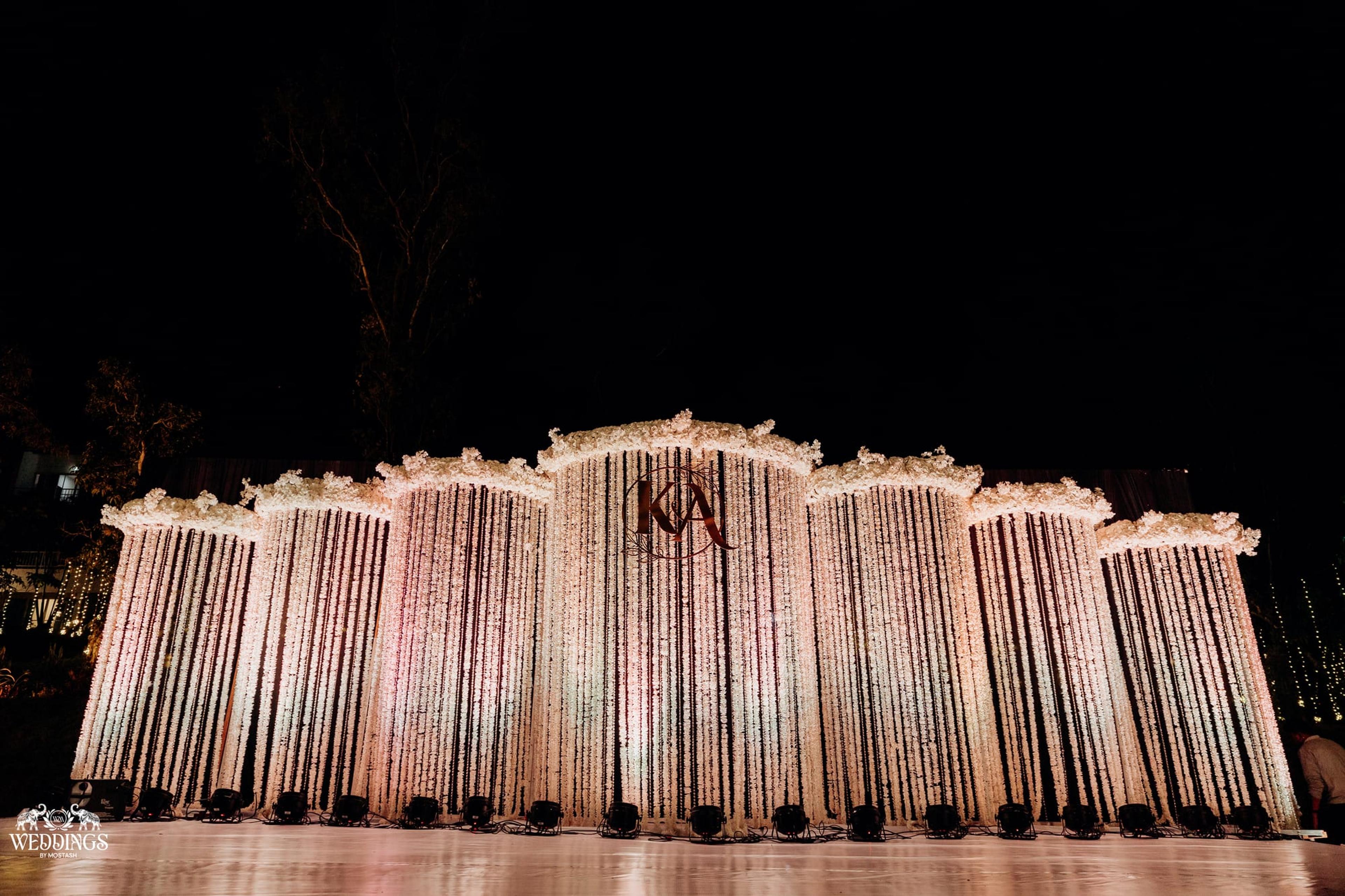 A grand reception stage with a floral-strand curtain backdrop and gold monogram washed in cool blue spotlight beams