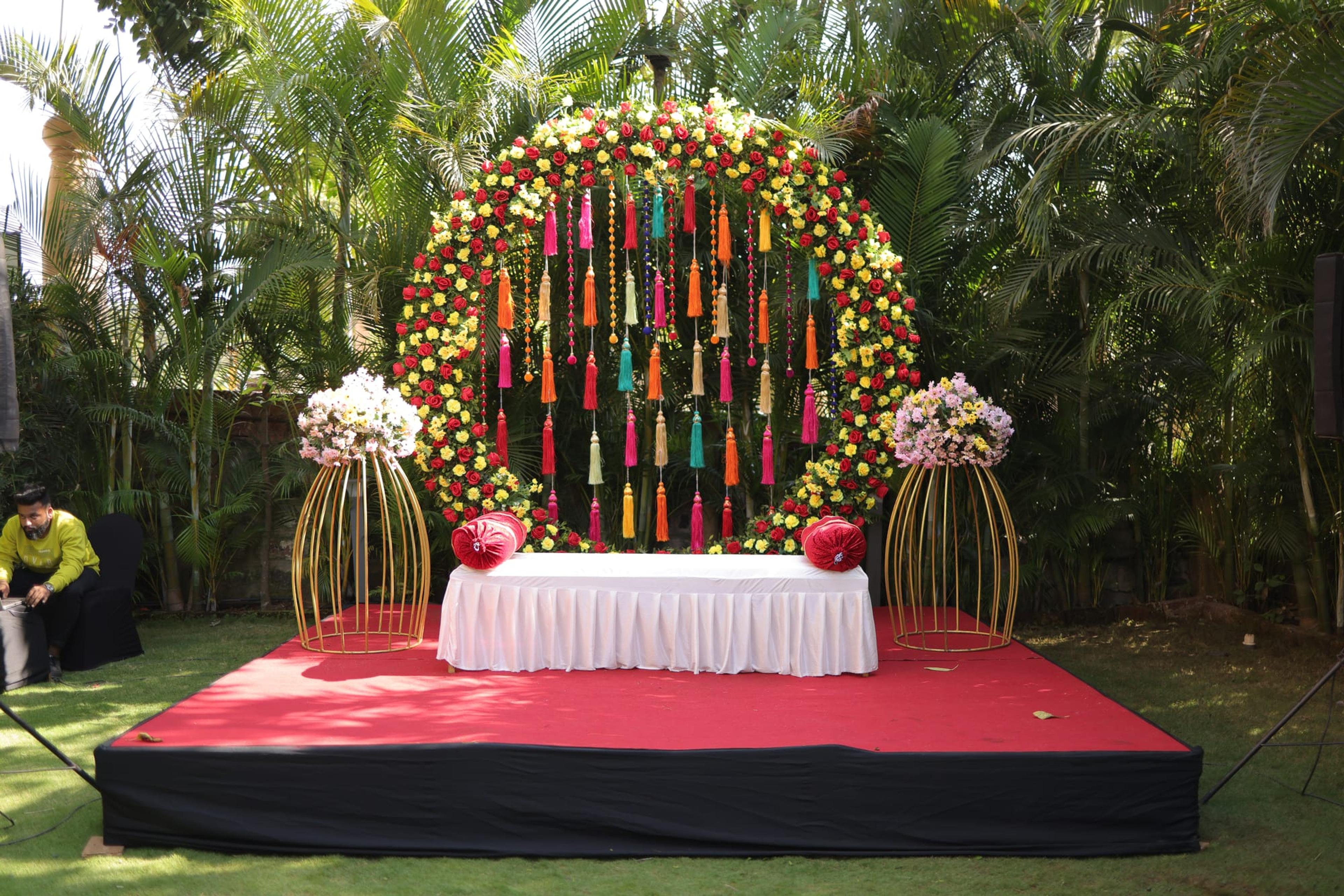 Circular marigold-and-rose floral arch hung with rainbow tassels above a red stage, flanked by gold birdcage flower stands