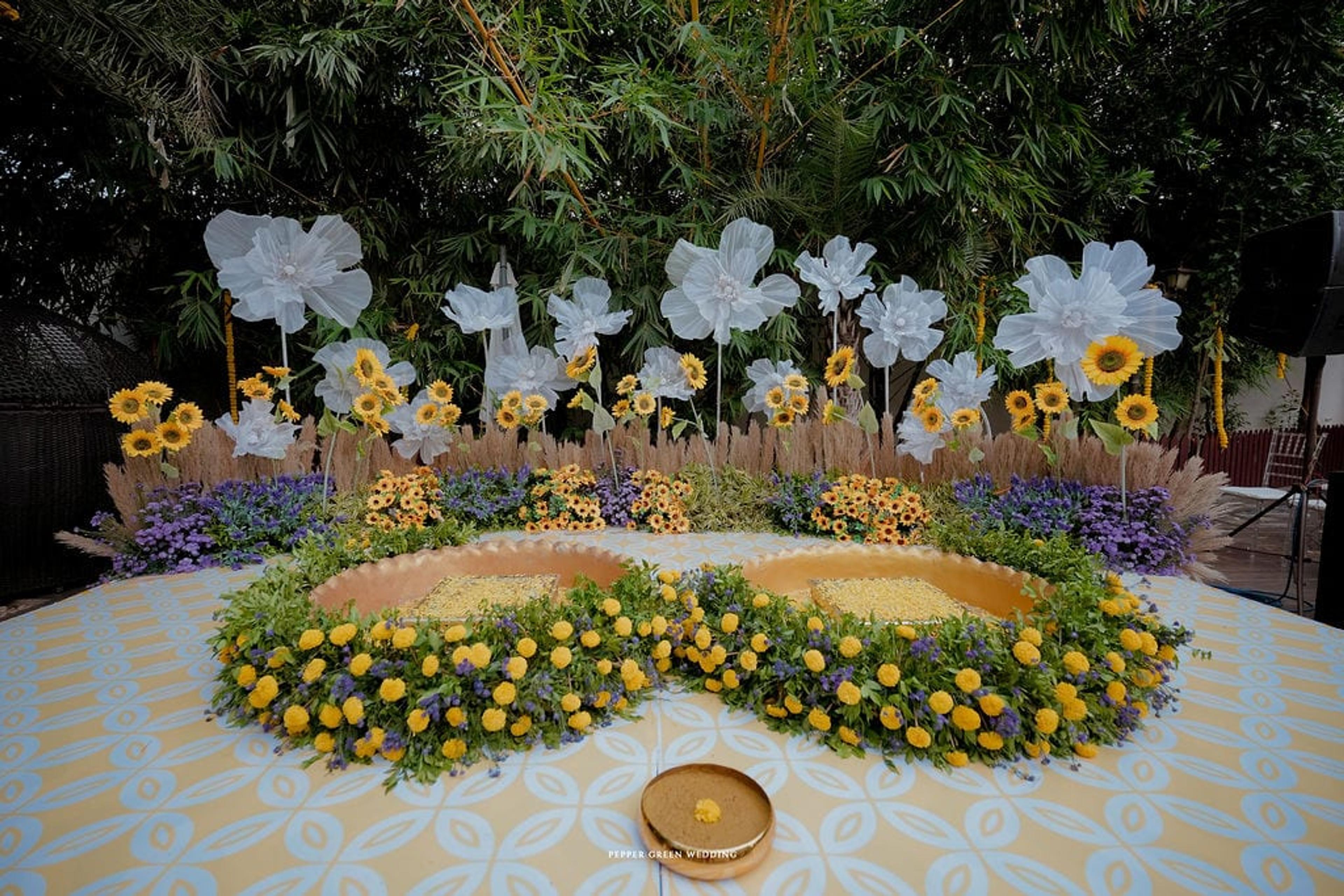 Marigold and white floral ceremony stage with a circular yellow-petal urli on a palm-lined resort lawn
