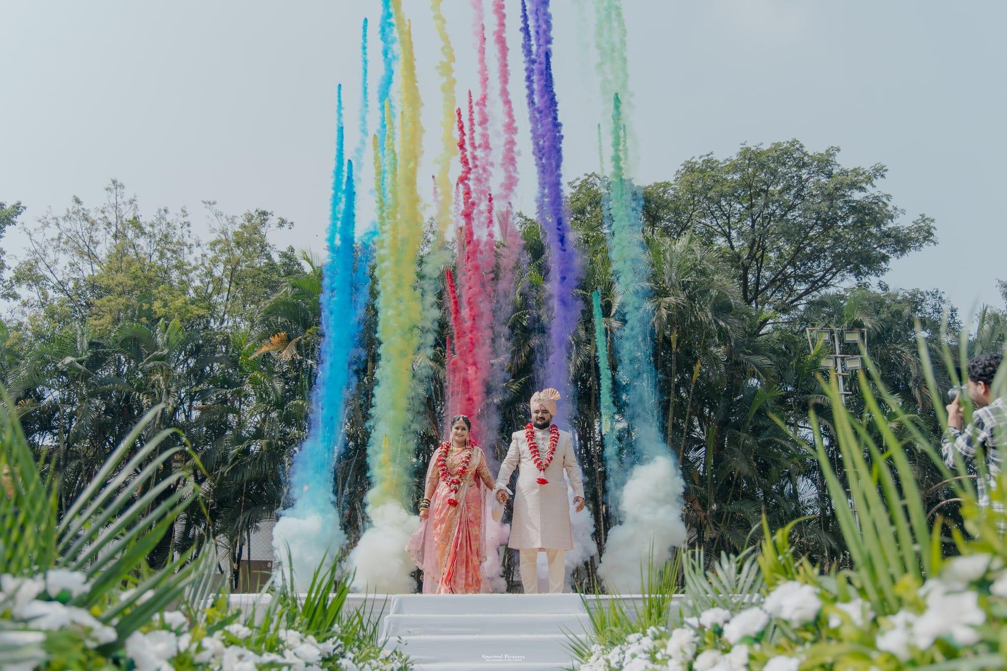 A couple walking through jets of rainbow colour-smoke in a sunlit garden