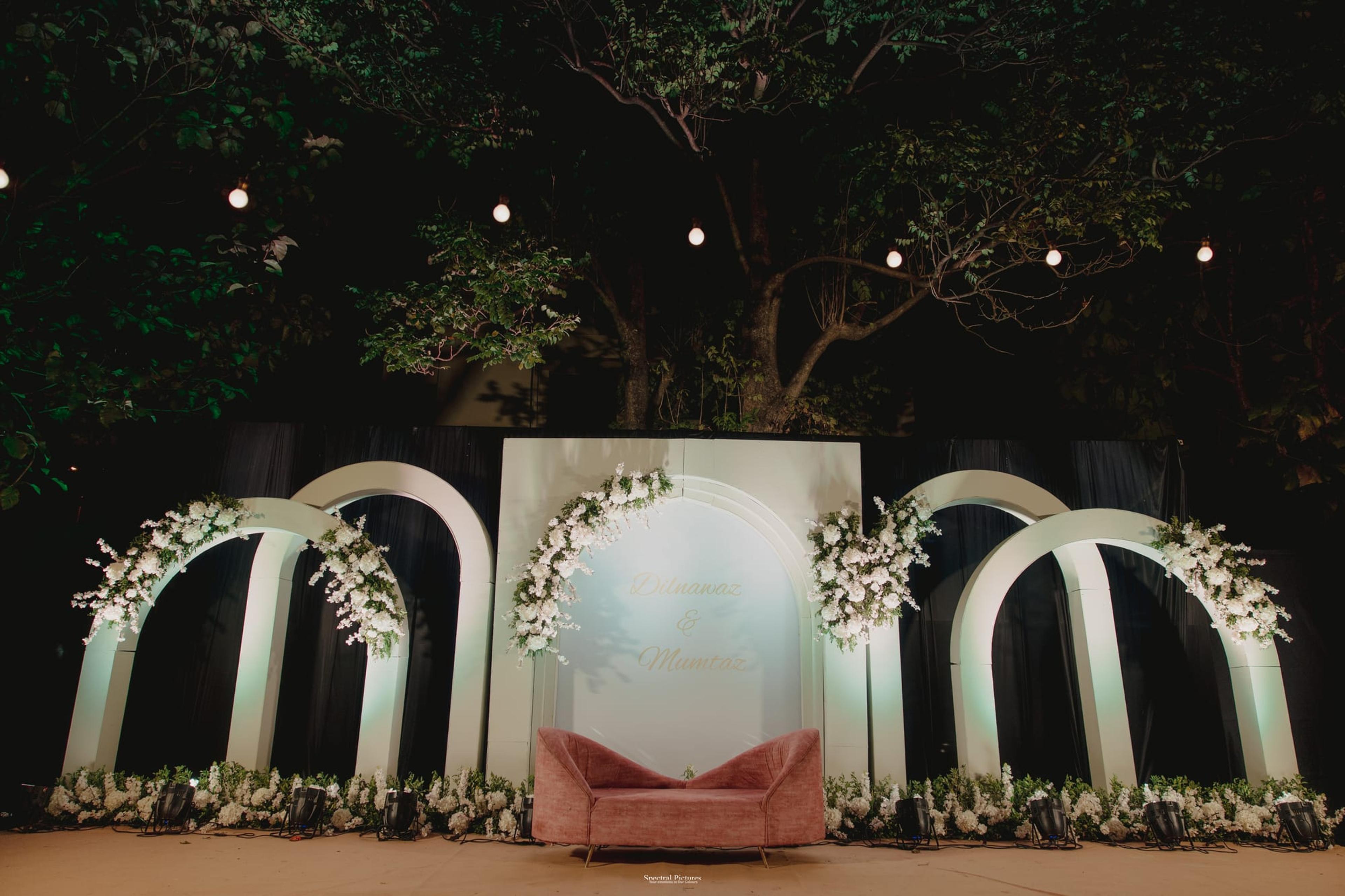 A floral white arch engagement stage with a pink platform set against lush green foliage and warm fairy lights