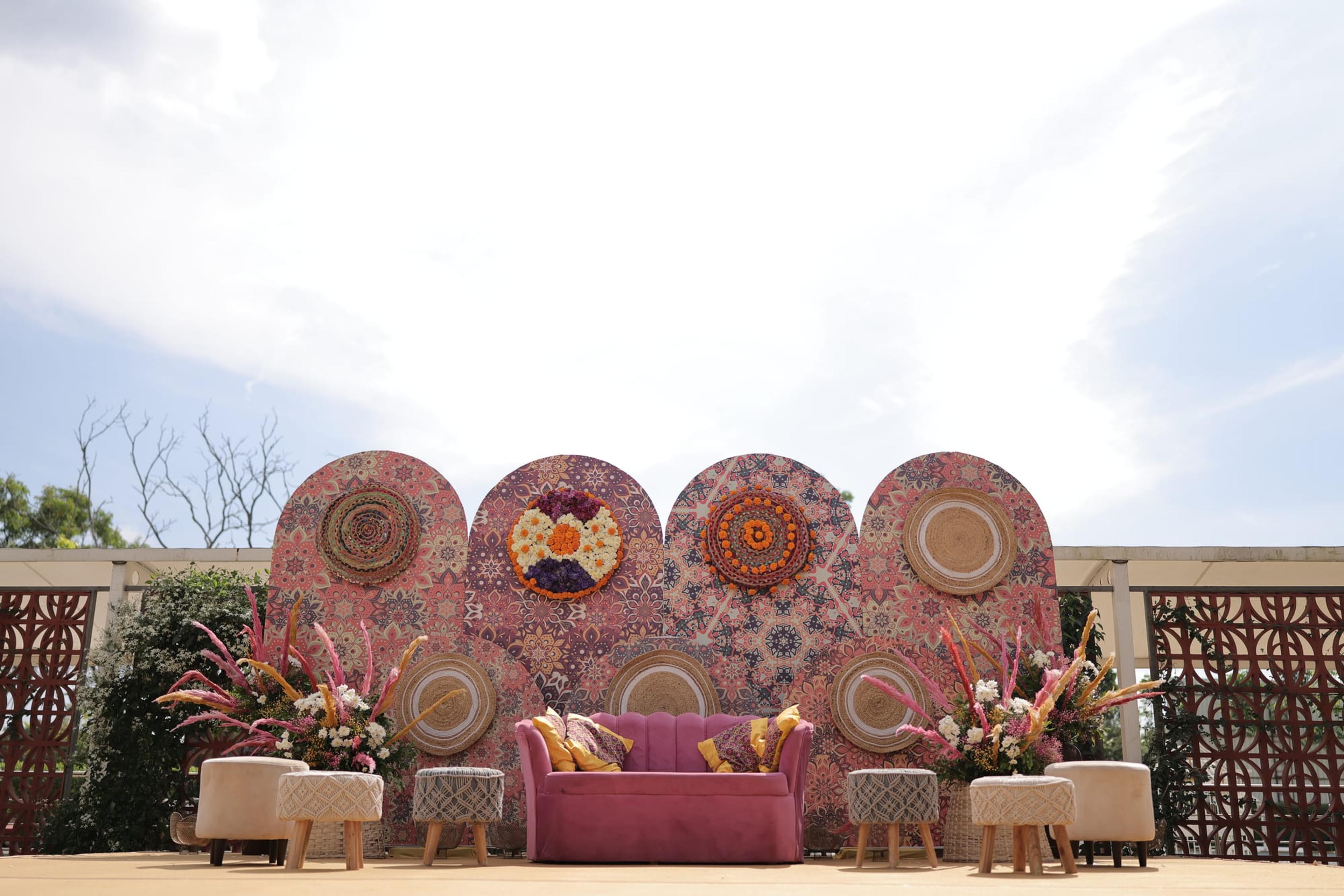 An outdoor wedding stage backed by oversized floral mandala discs in orange, pink and gold with a magenta sofa