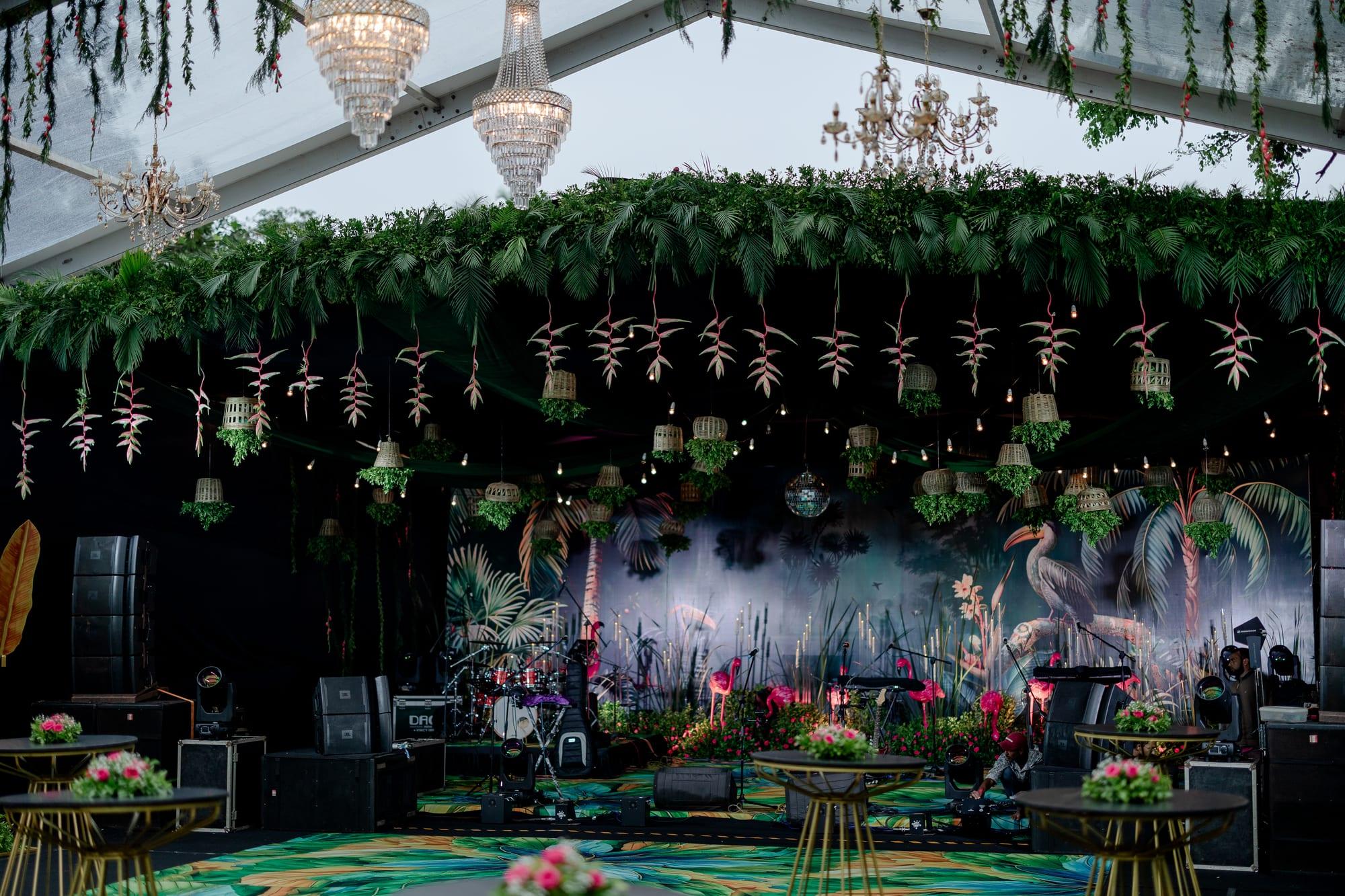 Tropical glasshouse stage under a thick canopy of hanging greenery and crystal chandeliers, with pink flamingo sculptures