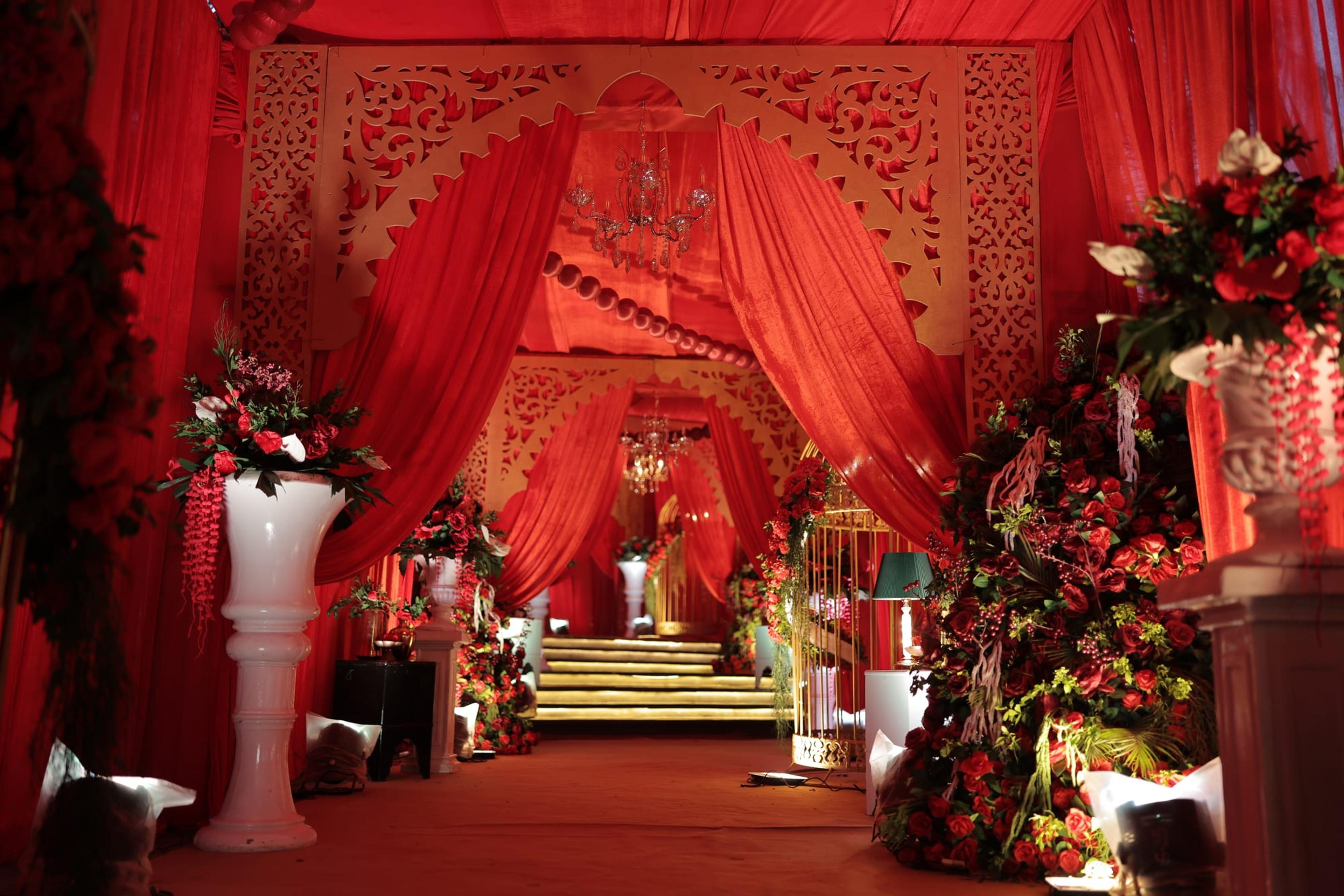 Ornate carved ivory nikah stage draped in crimson fabric with a crystal chandelier and towering red rose floral pillars