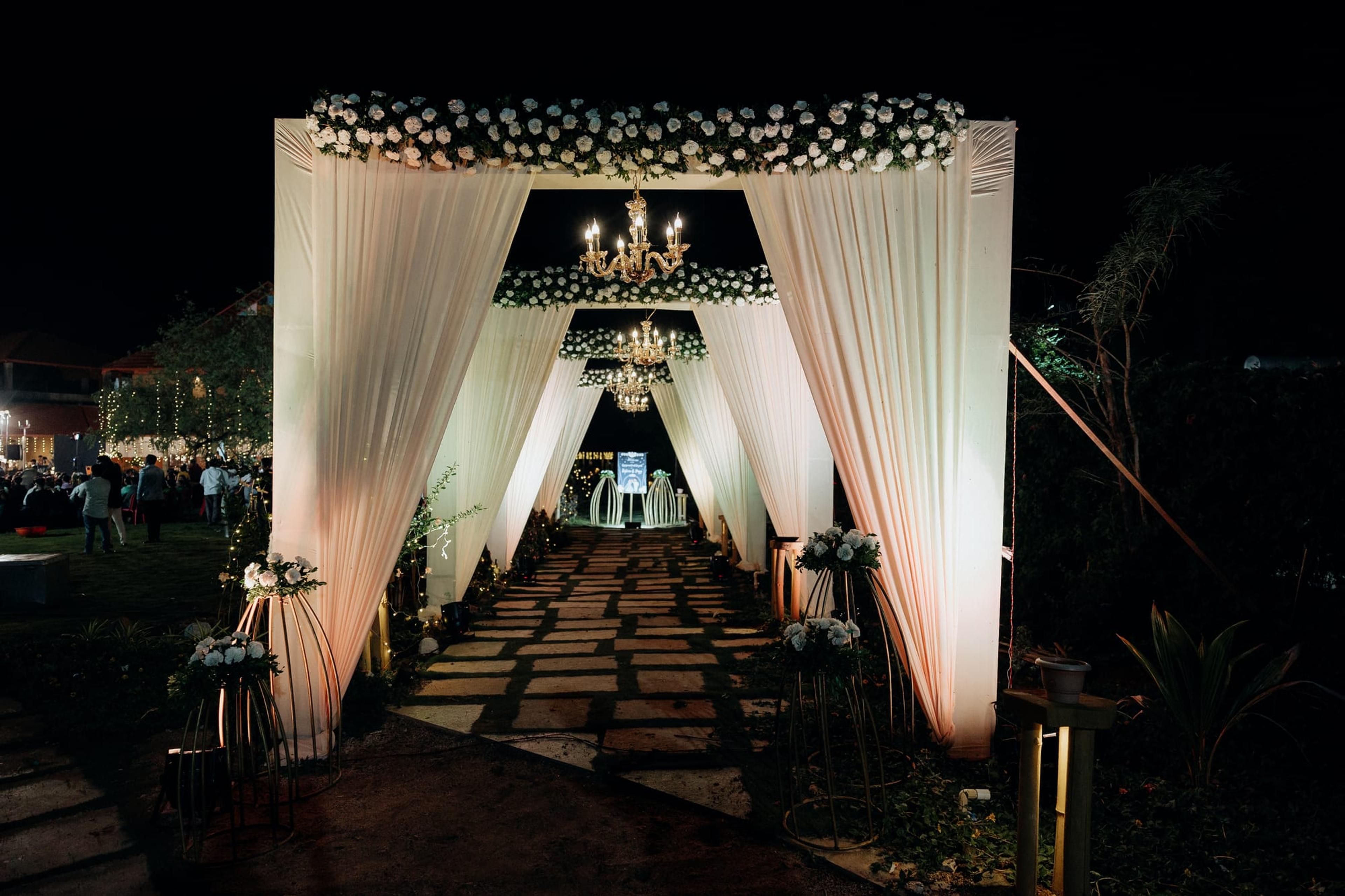 A draped, candlelit heritage entrance walkway hung with chandeliers leading to a glowing floral stage