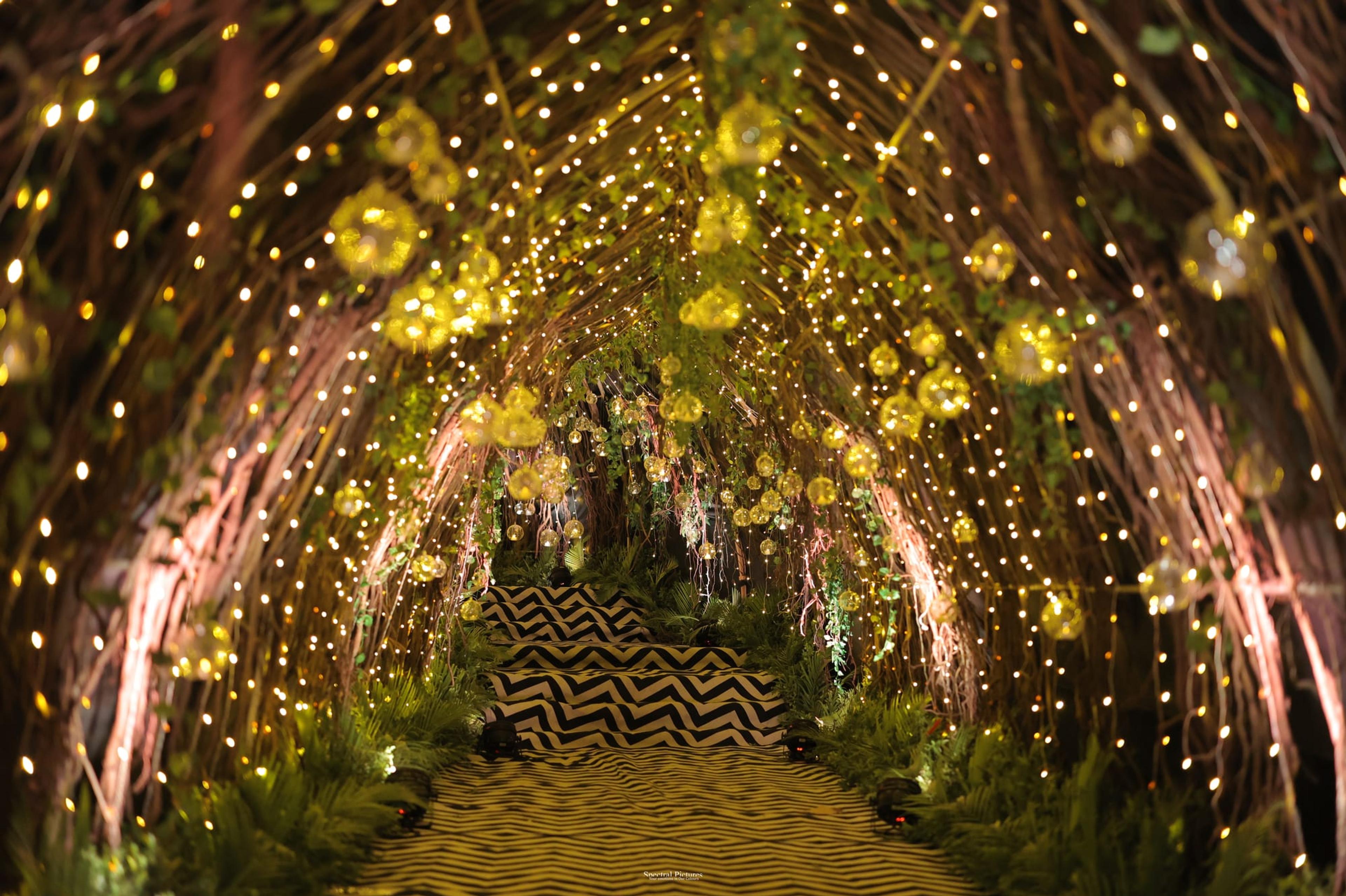 A fairy-light floral entrance tunnel of arched greenery glowing with warm amber light over a candle-lined garden aisle