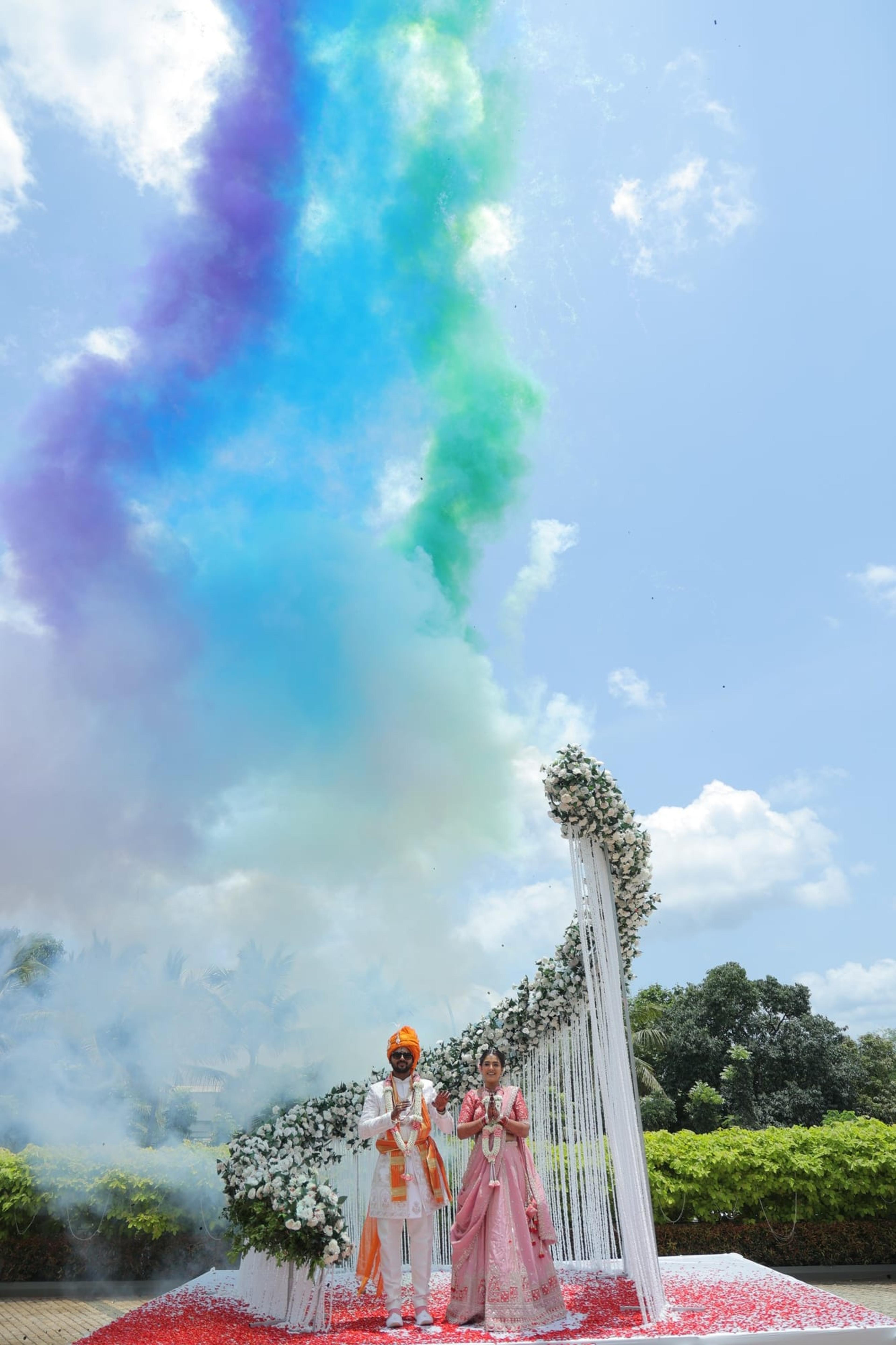 Bride and groom beneath a towering white-floral mandap as a rainbow plume of colour-smoke erupts over a rose-petal carpet