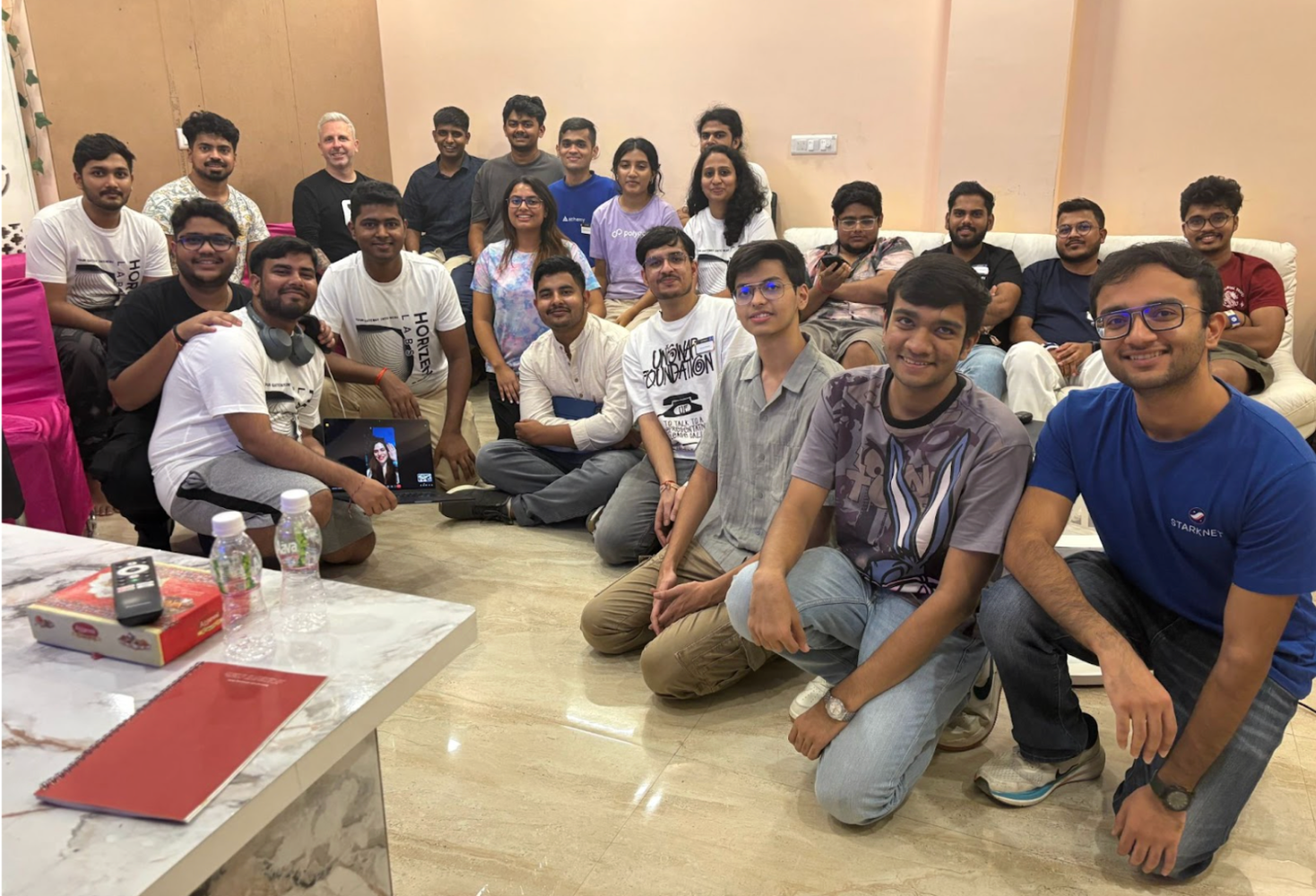 A group of young adults gathered in a casual room, some on a couch, many on the floor, with a laptop displaying a video call.