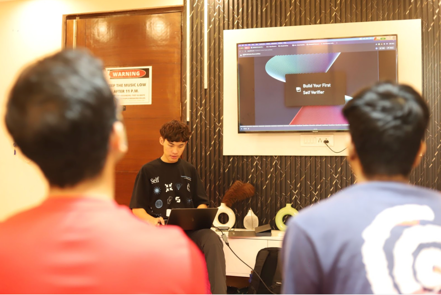 A young man uses a laptop while presenting to two people, with a large screen displaying "Build Your First Self Verifier."
