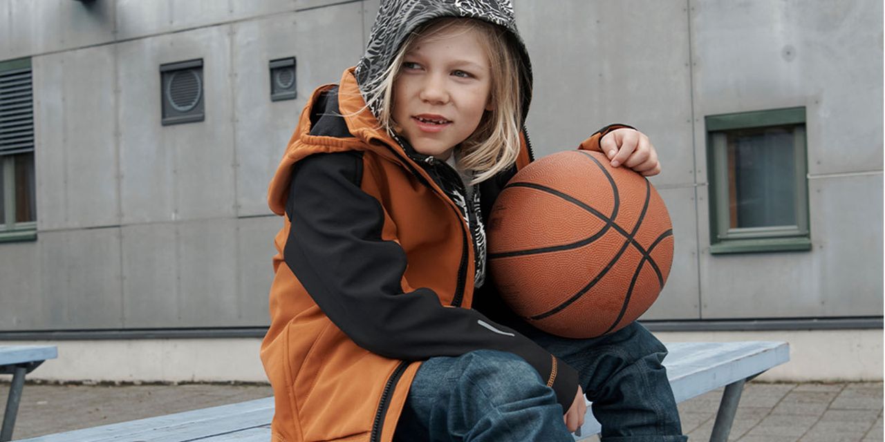 Ett äldre barn som sitter på en bänk, med en basketboll i famnen. Barnet bär en orange och svart skaljacka och har luvan på sig.