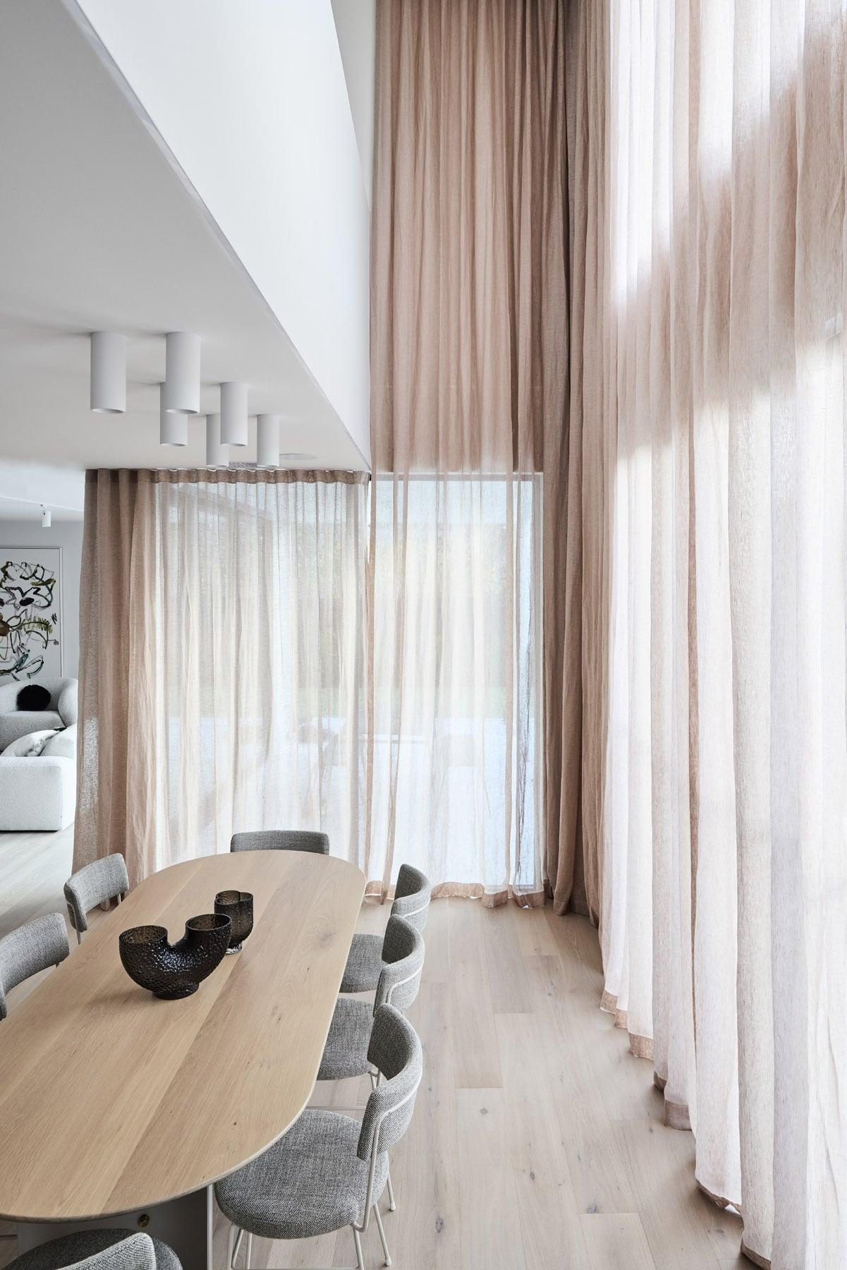 Thomas Archer Architecturally Designed Elba Display Home modern dining area with a wooden table, grey chairs, and sheer floor-to-ceiling curtains. 