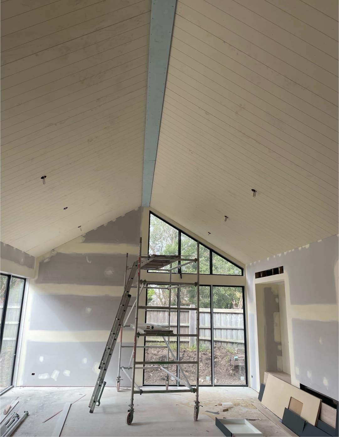 A room with a ceiling being painted. The painter is standing on a ladder, applying paint to the ceiling surface.