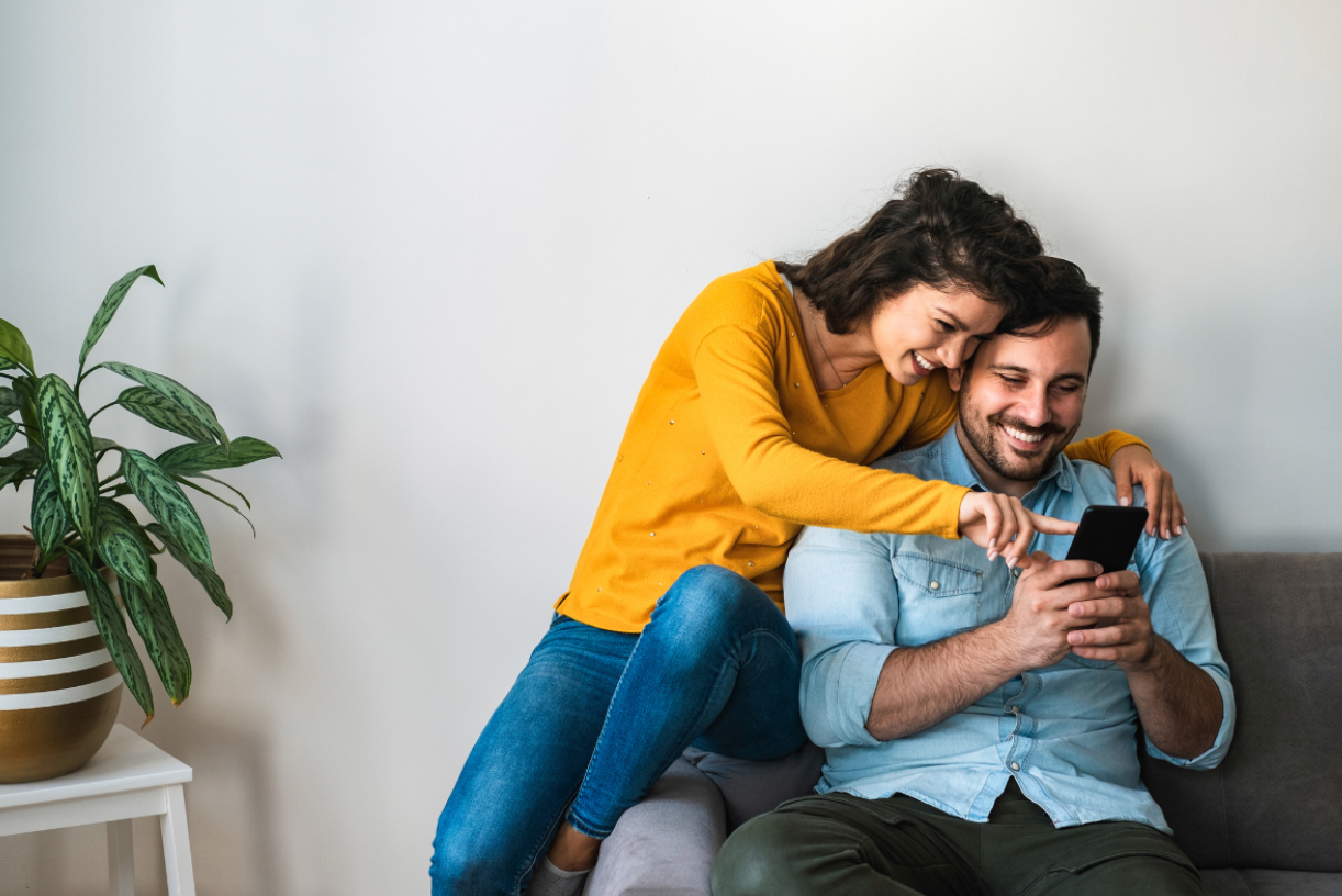 A couple sitting on a couch, smiling, and looking at the phone.