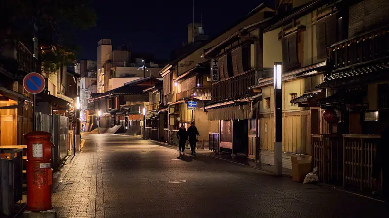 Two pedestrians on a quiet street lined with traditional buildings in Gion, Kyoto at night