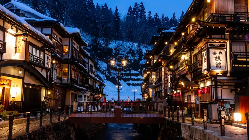 Snow-covered ryokan and illuminated lamps along the river at dusk in Ginzan Onsen, Yamagata