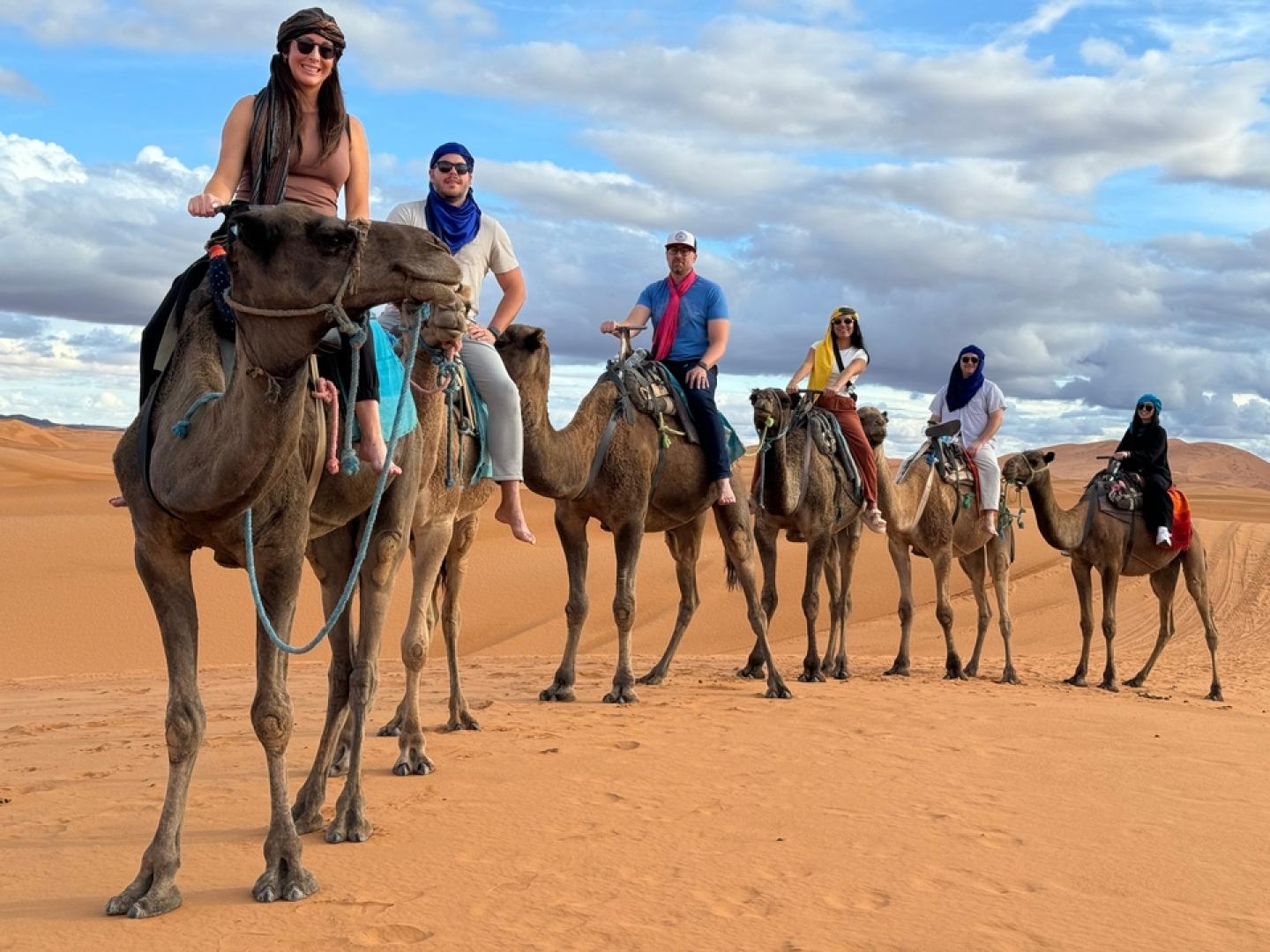 Six guests in colourful desert turbans and sunglasses lined up on camels in the Sahara dunes