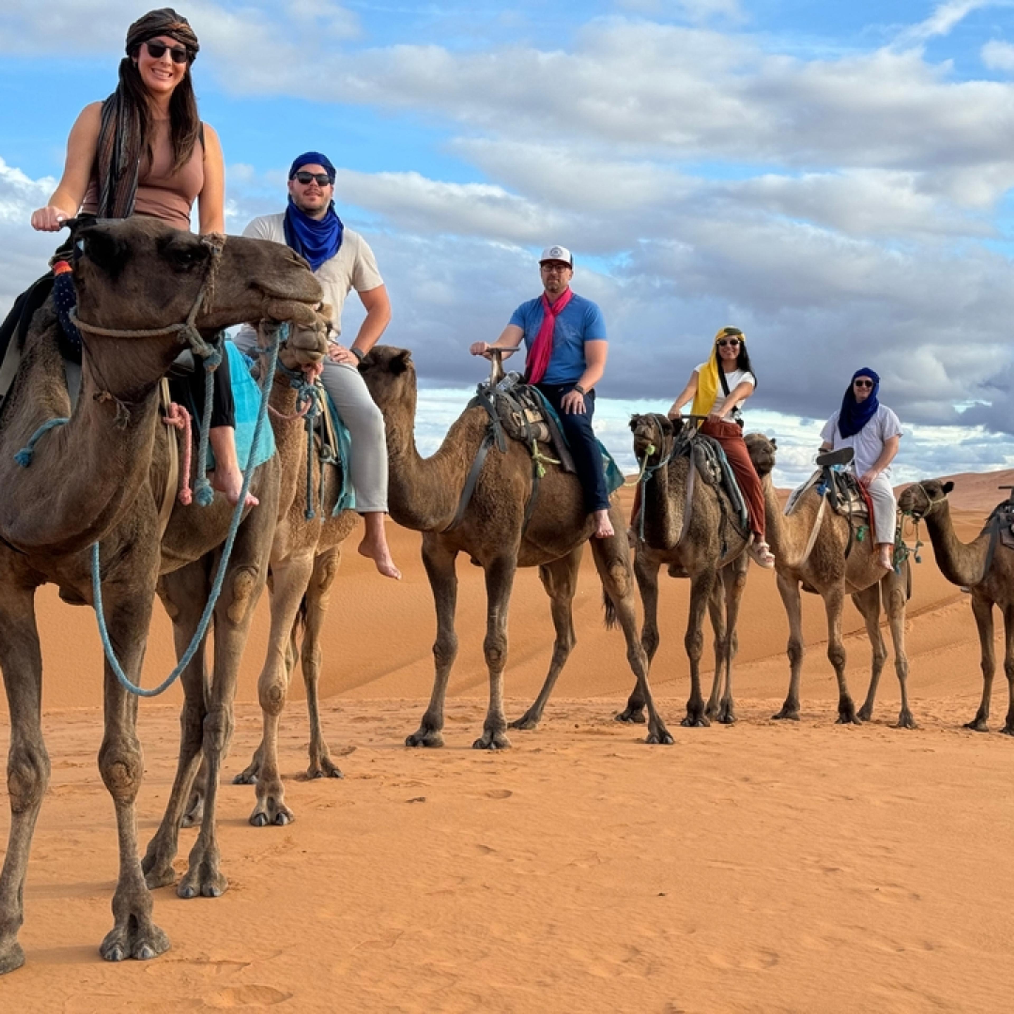 Six guests in colourful desert turbans and sunglasses lined up on camels in the Sahara dunes