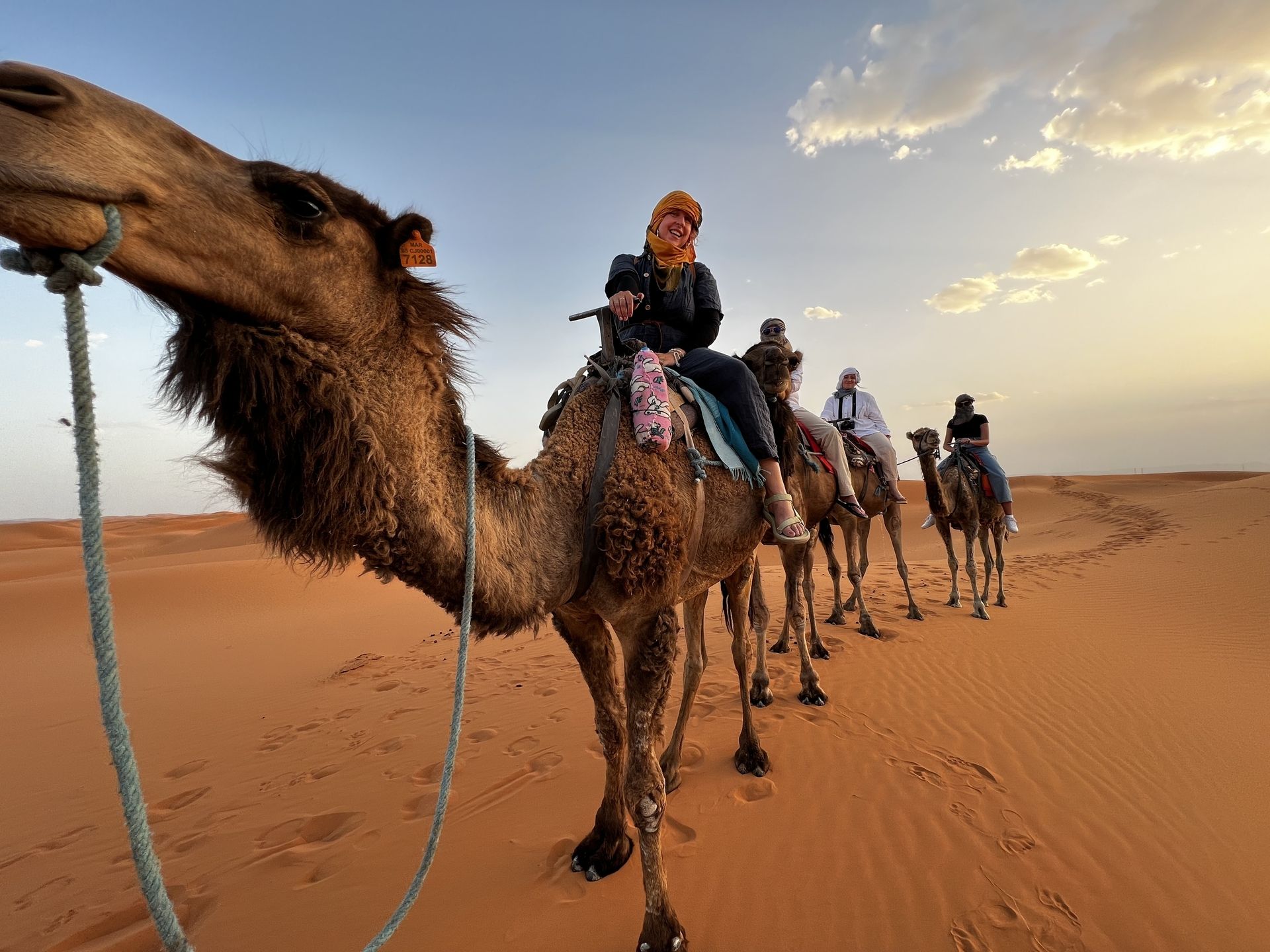 A smiling guest in an orange headscarf on a camel at sunset, with three more riders following across the dunes