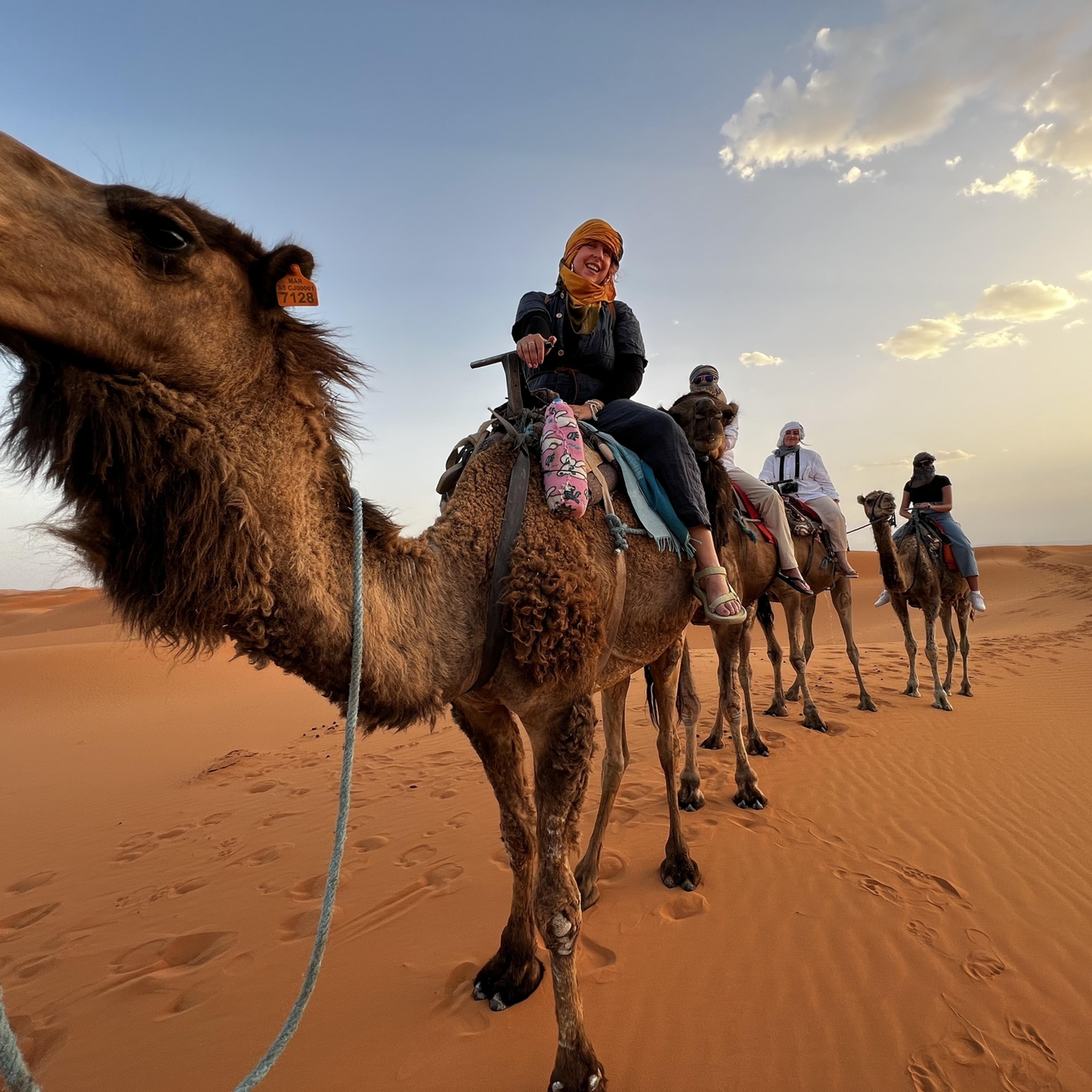 A smiling guest in an orange headscarf on a camel at sunset, with three more riders following across the dunes