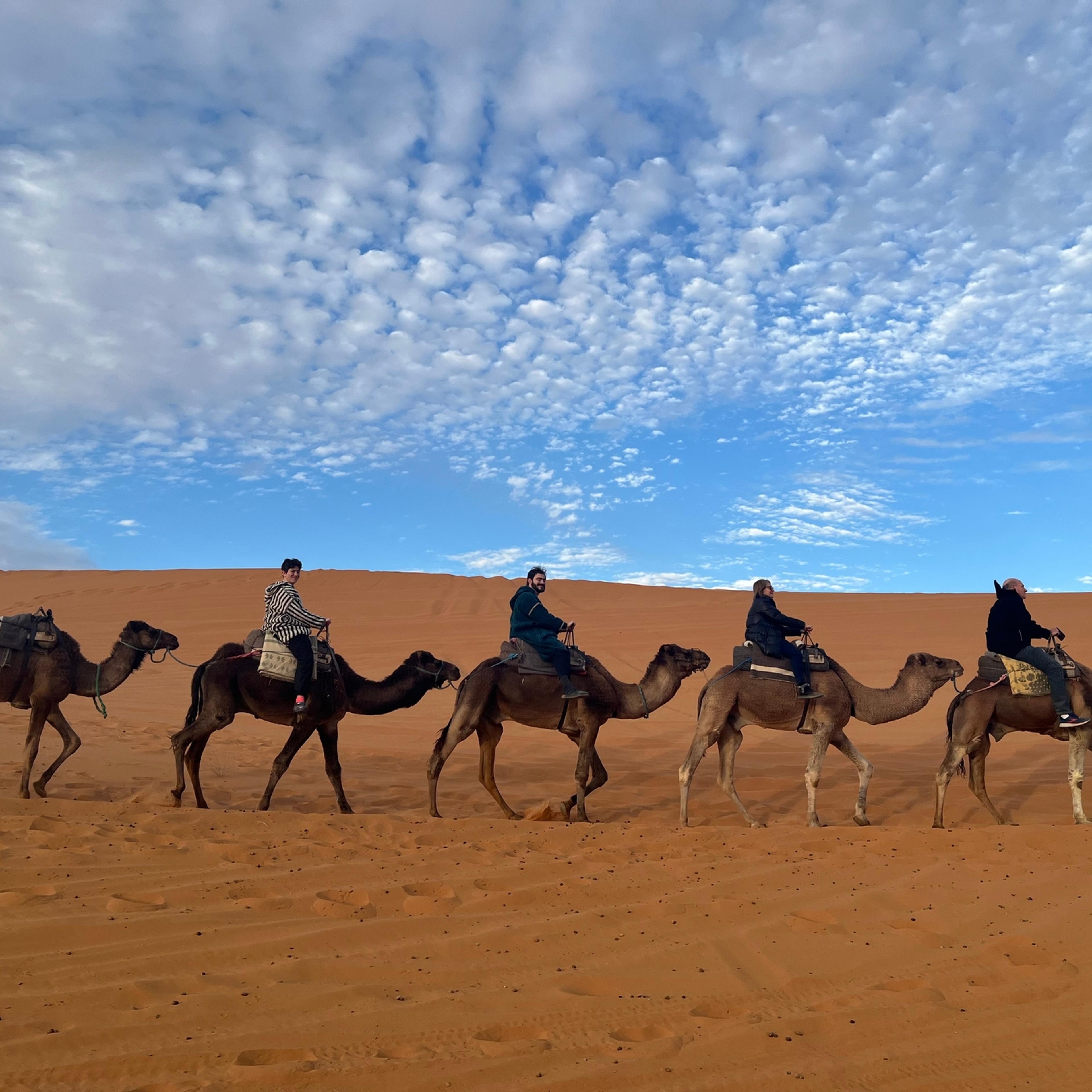 Four guests riding a camel caravan along a ridge of orange Sahara dunes under a sky full of small clouds