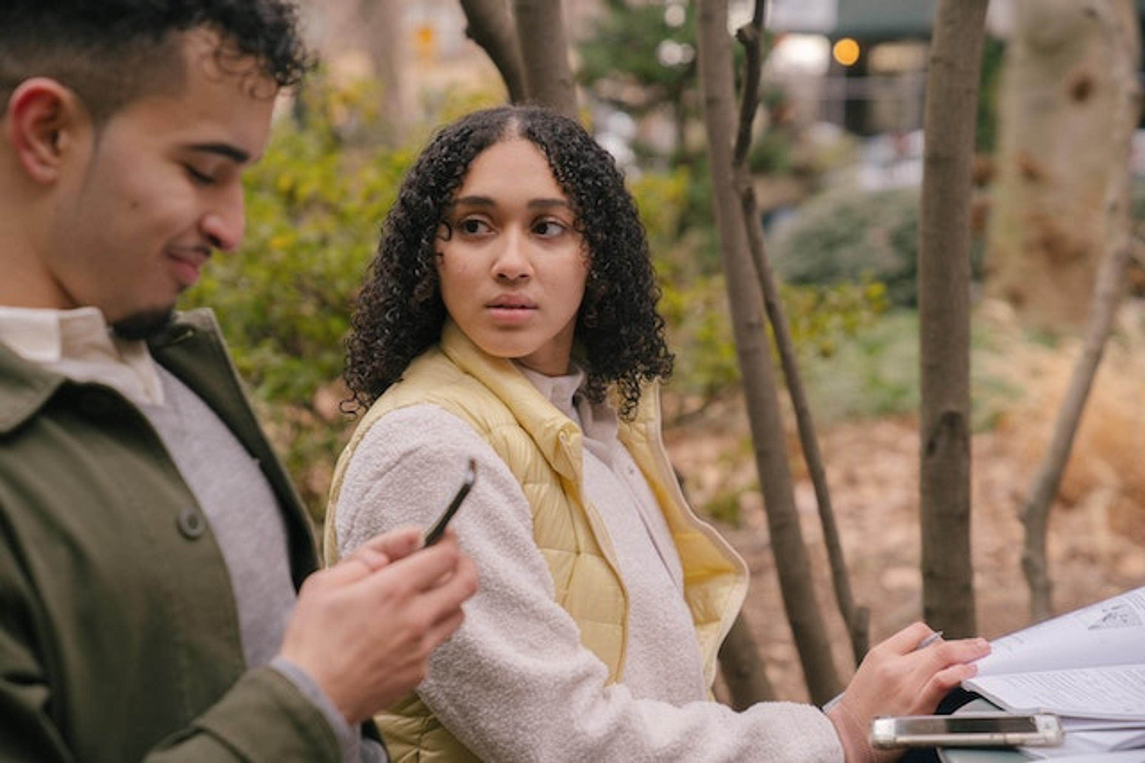 A man and a woman are sitting outside where the man is sitting with his phone. The women is feeling retroactive realousy.